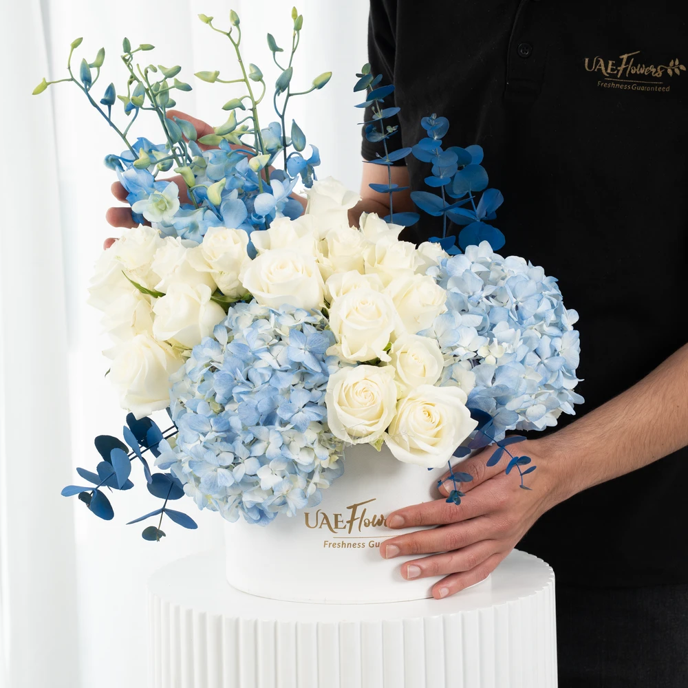 A box arrangement of 2 blue hydrangeas, 20 white roses, 5 blue orchids and blue sprayed eucalyptus leaf.