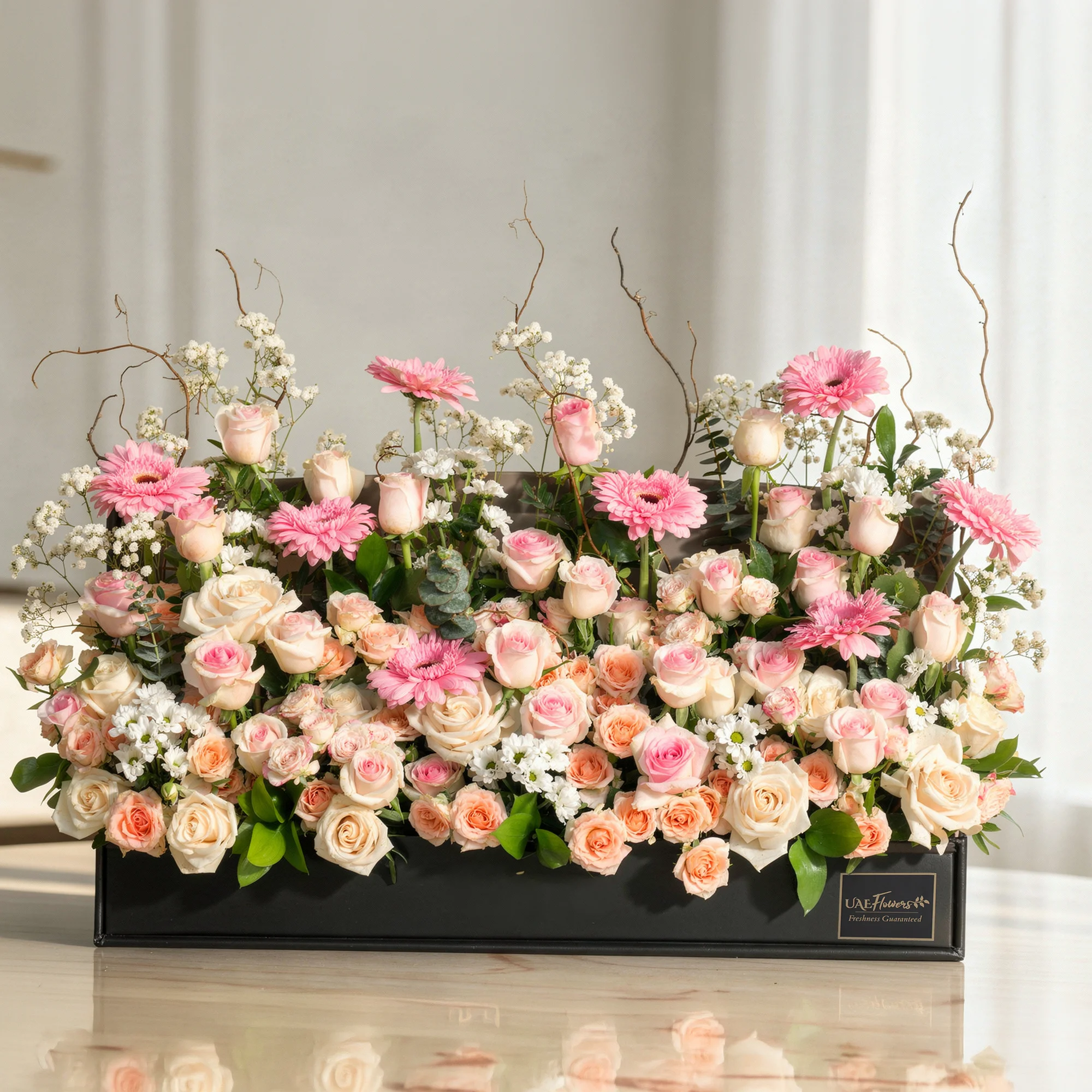 Arrangement of 8 pink gerberas, 30 pink roses, 10 peach roses, 10 Pink spray roses and 5 white chrysanthemums with green fillers and baby's breath in a long black box.