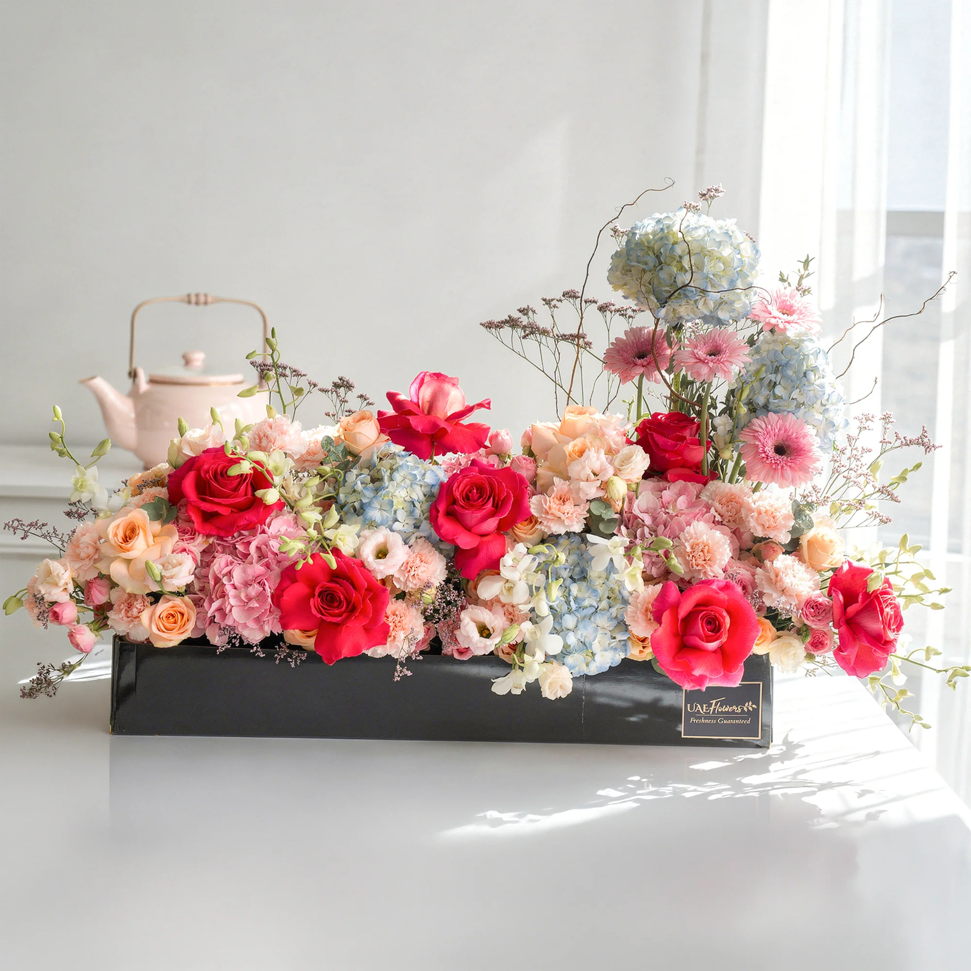 Arrangement of 4 blue hydrangeas, 2 pink hydrangeas, 20 pink carnations, 10 pink spray roses, 10 peach roses and 4 pink gerberas with green fillers in a long black box.