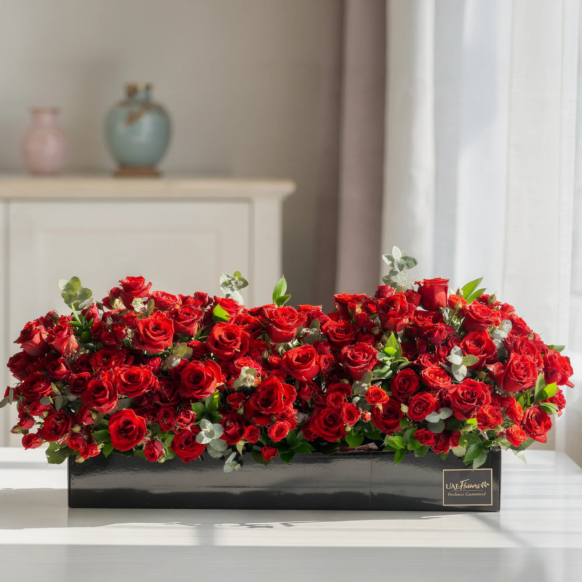 Arrangement of 50 red roses and 30 red spray roses with green fillers in a long black box.