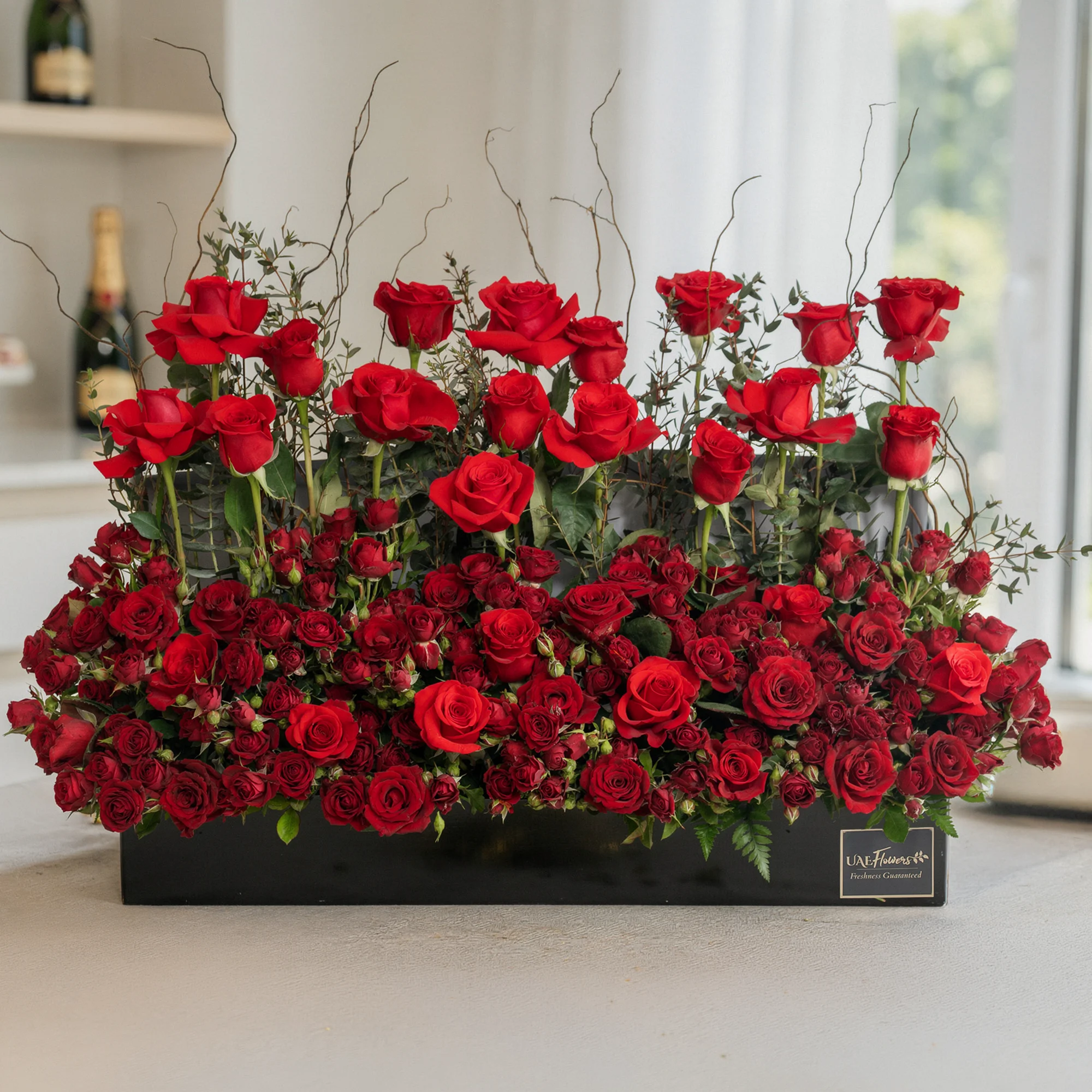 Arrangement of 50 red roses and 20 red spray roses with green fillers in a long black box.