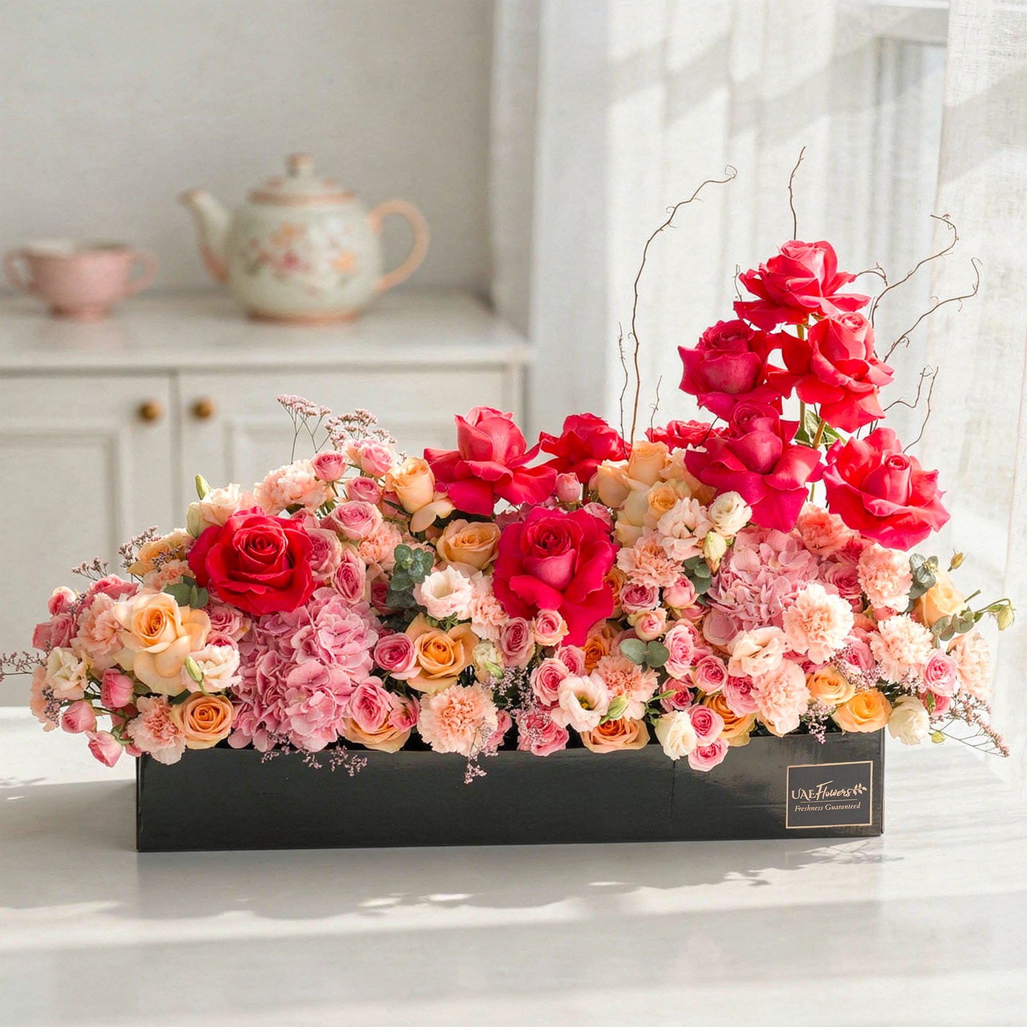 Arrangement of 10 fuchsia roses, 20 pink carnations, 20 pink spray roses, 20 peach roses and 2 pink hydrangeas with green fillers in a long black box.