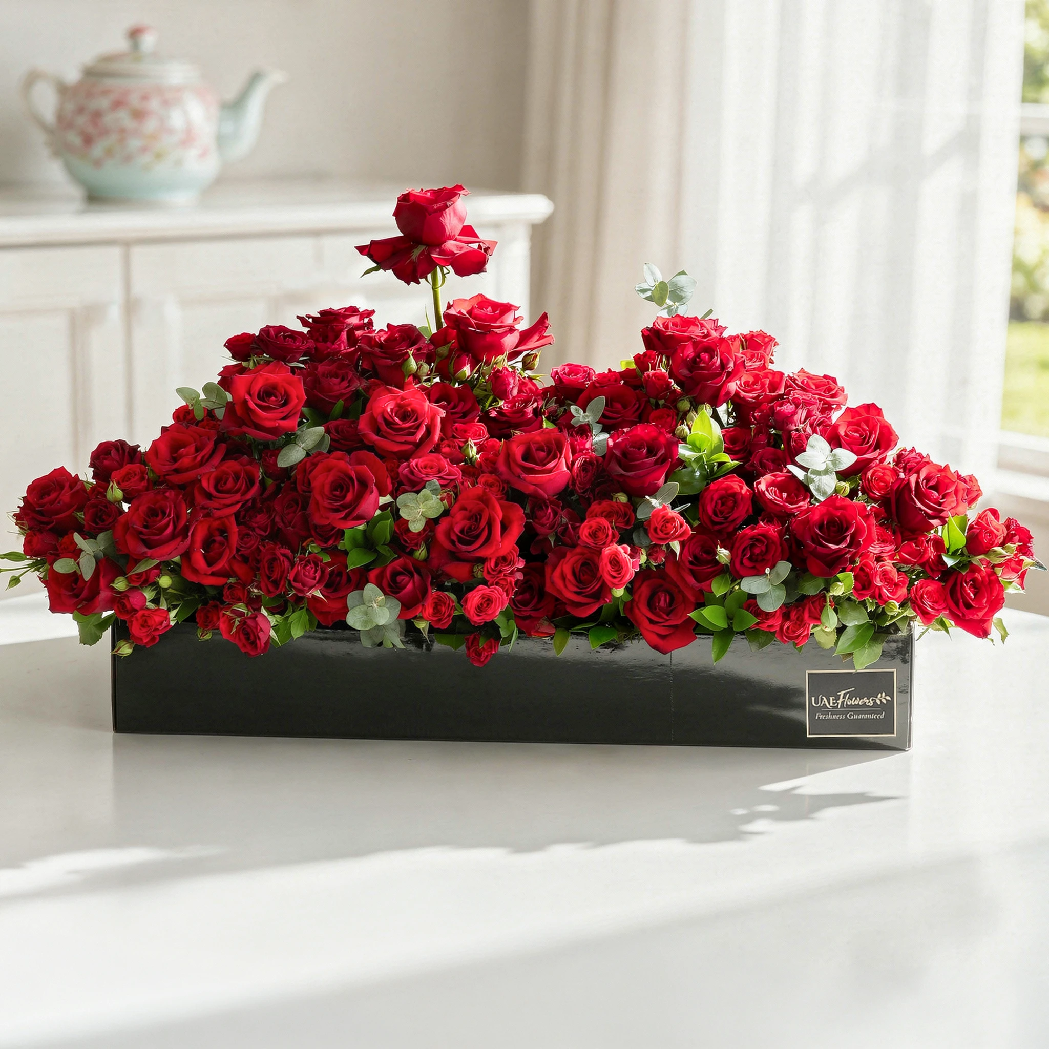 Arrangement of 50 red roses and 30 red spray roses with green fillers in a long black box.
