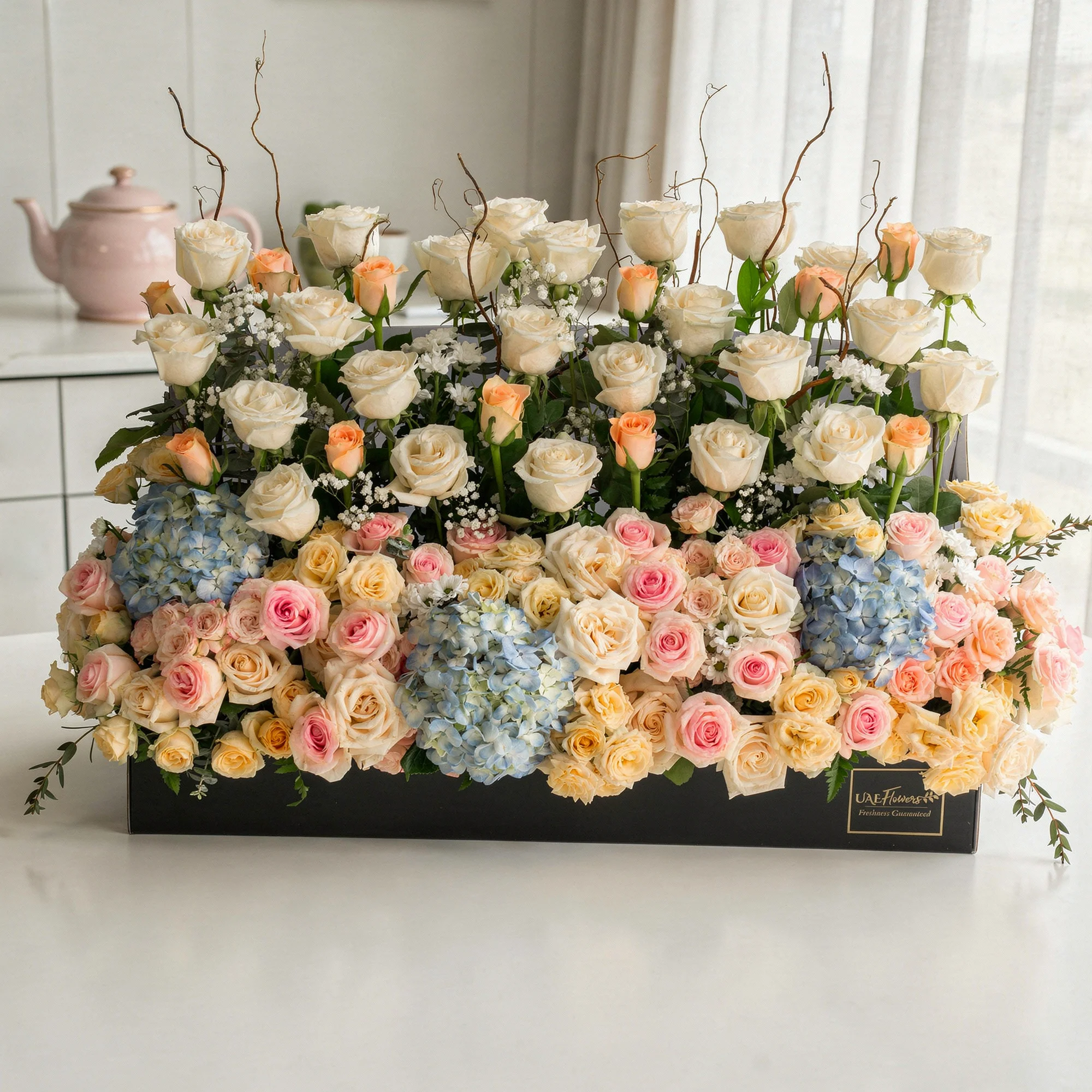 Arrangement of 30 White roses, 20 pink roses, 20 peach roses, 3 blue hydrangeas and 10 peach spray roses with green fillers in a long black box.