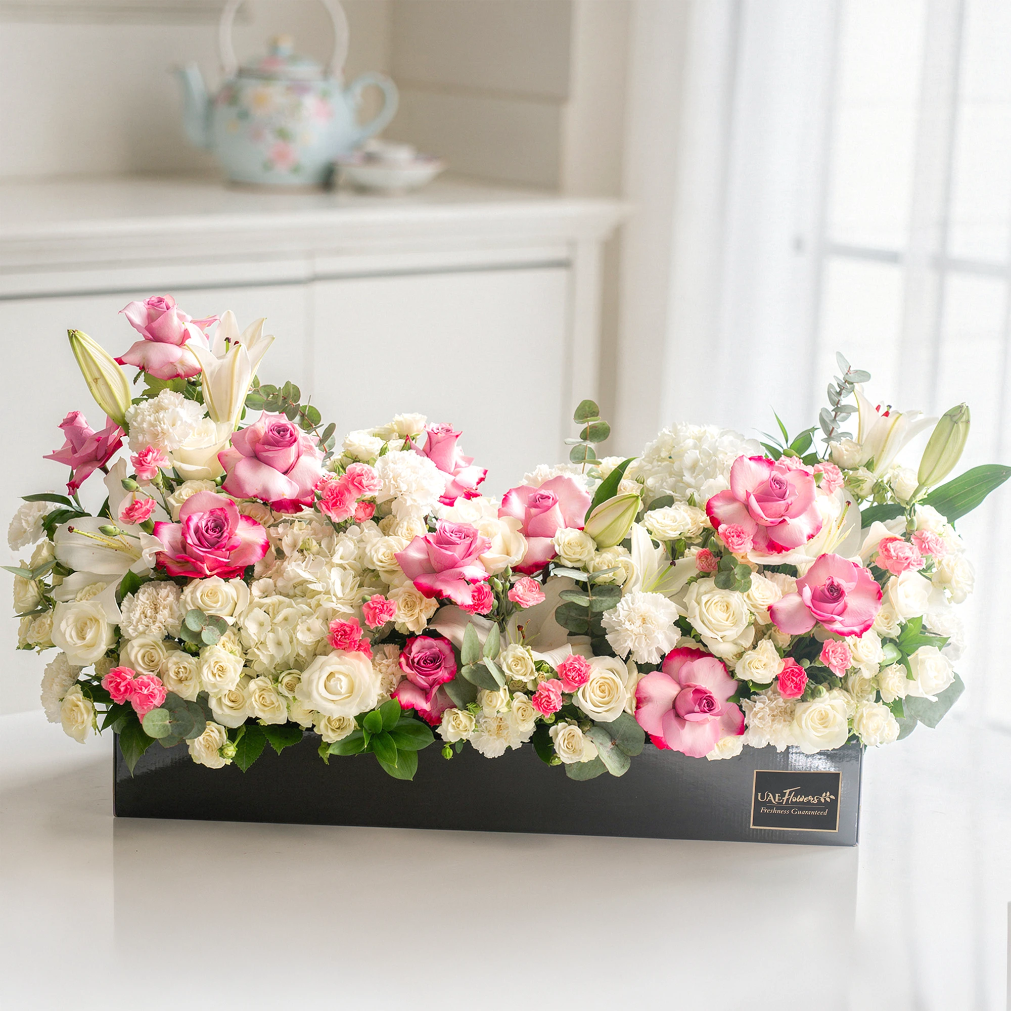 Arrangement of 5 white lilies, 15 purple roses, 15 white roses, 10 white carnations, 10 pink carnations, 10 white spray roses and 2 white hydrangeas with green fillers in a long black box.