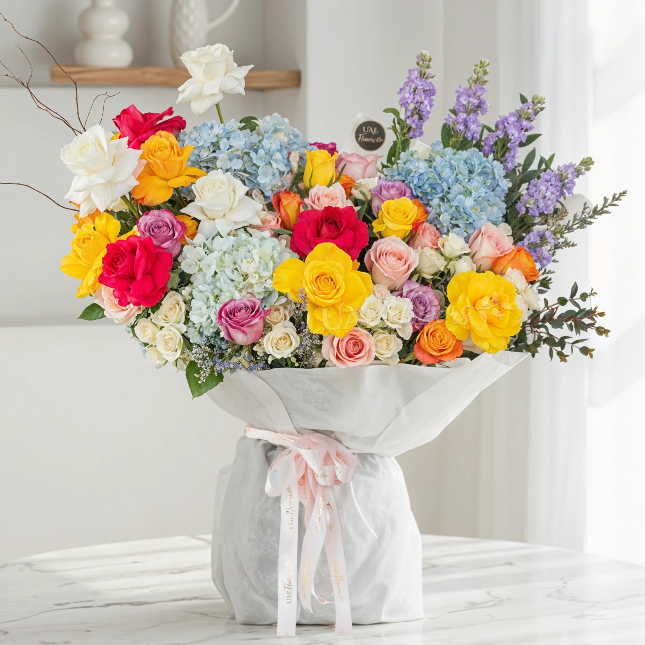 Bouquet of 25 mix roses, 6 white spray roses, 3 blue hydrangeas and 5 purple matthiola with green fillers in white paper packing tied with pink ribbon.