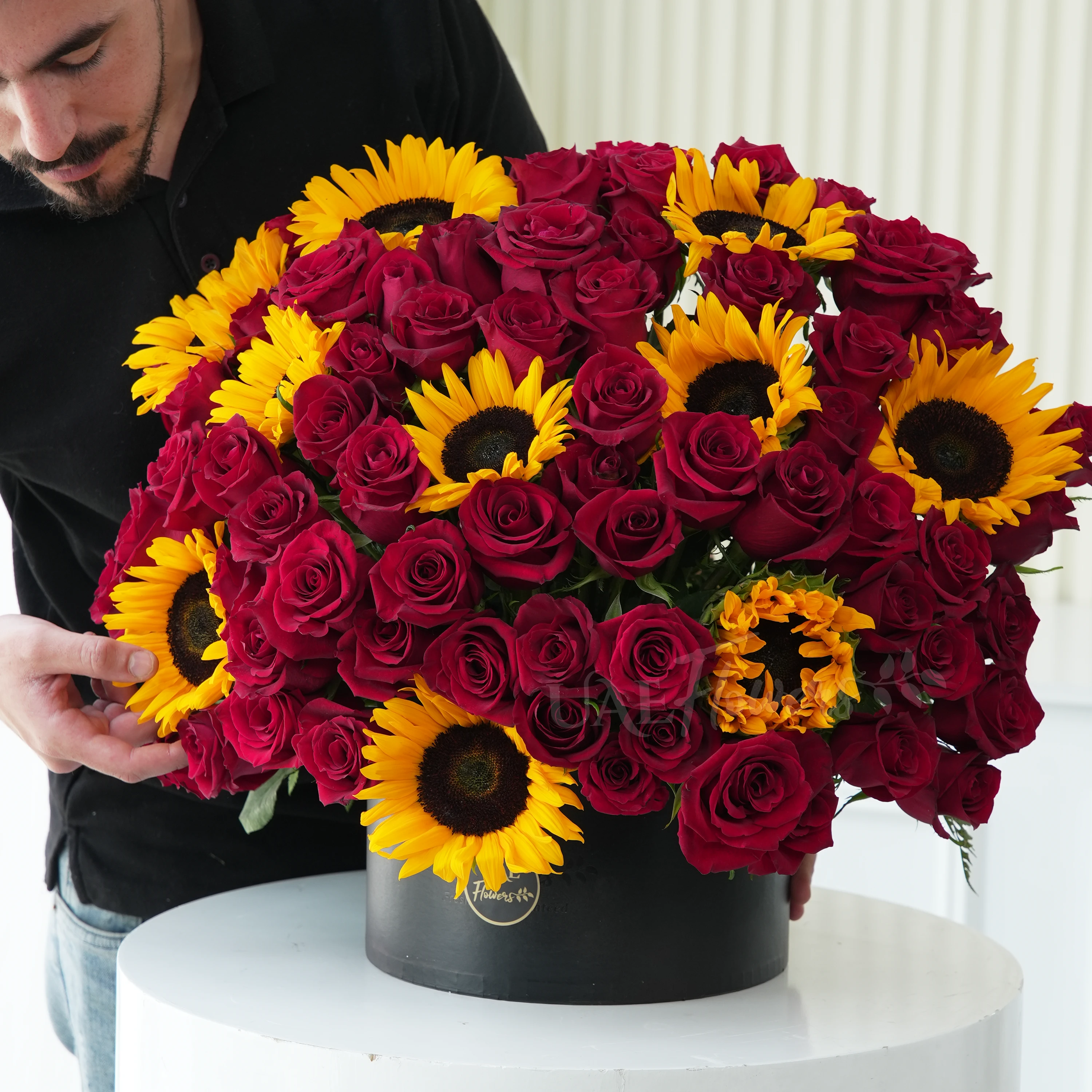 One side round arrangement of 10 sunflowers and 60 red roses with greens in a black round box.