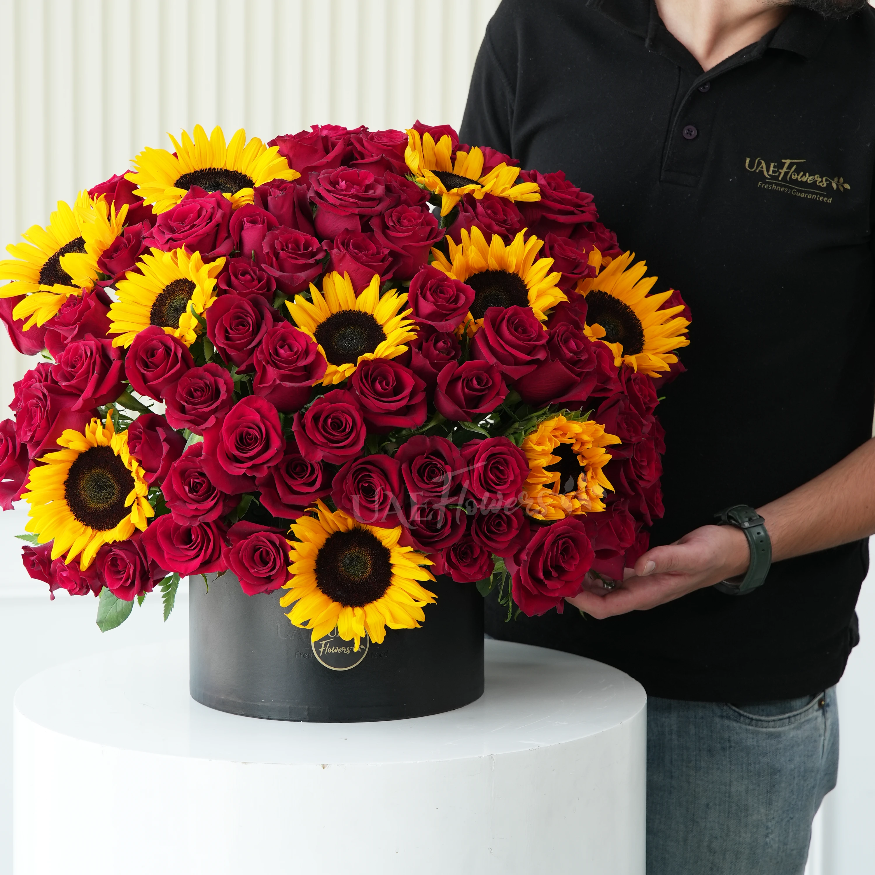 One side round arrangement of 10 sunflowers and 60 red roses with greens in a black round box.