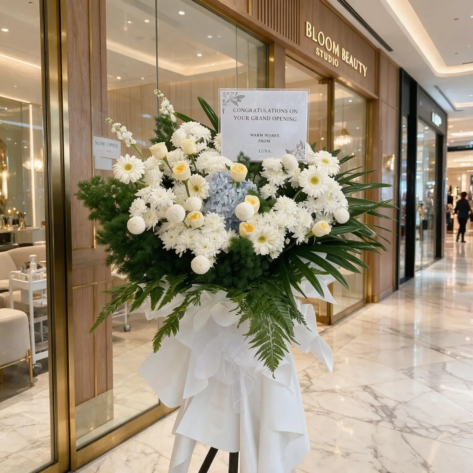 The Peaceful Serenity Sympathy Stand by UAE Flowers, featuring white chrysanthemums, peach roses, and blue hydrangea, presented with a condolence message card