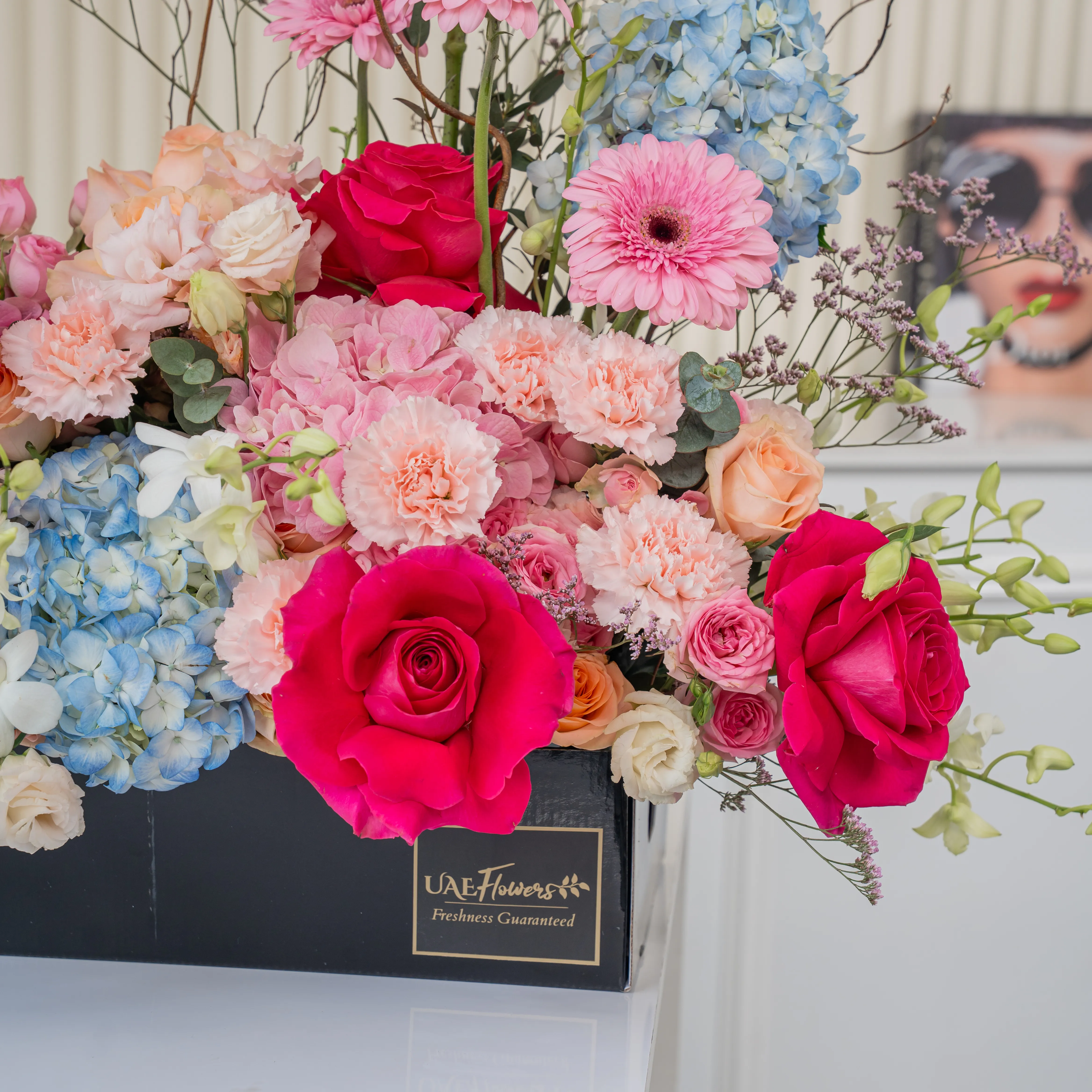 Arrangement of 4 blue hydrangeas, 2 pink hydrangeas, 20 pink carnations, 10 pink spray roses, 10 peach roses and 4 pink gerberas with green fillers in a long black box.