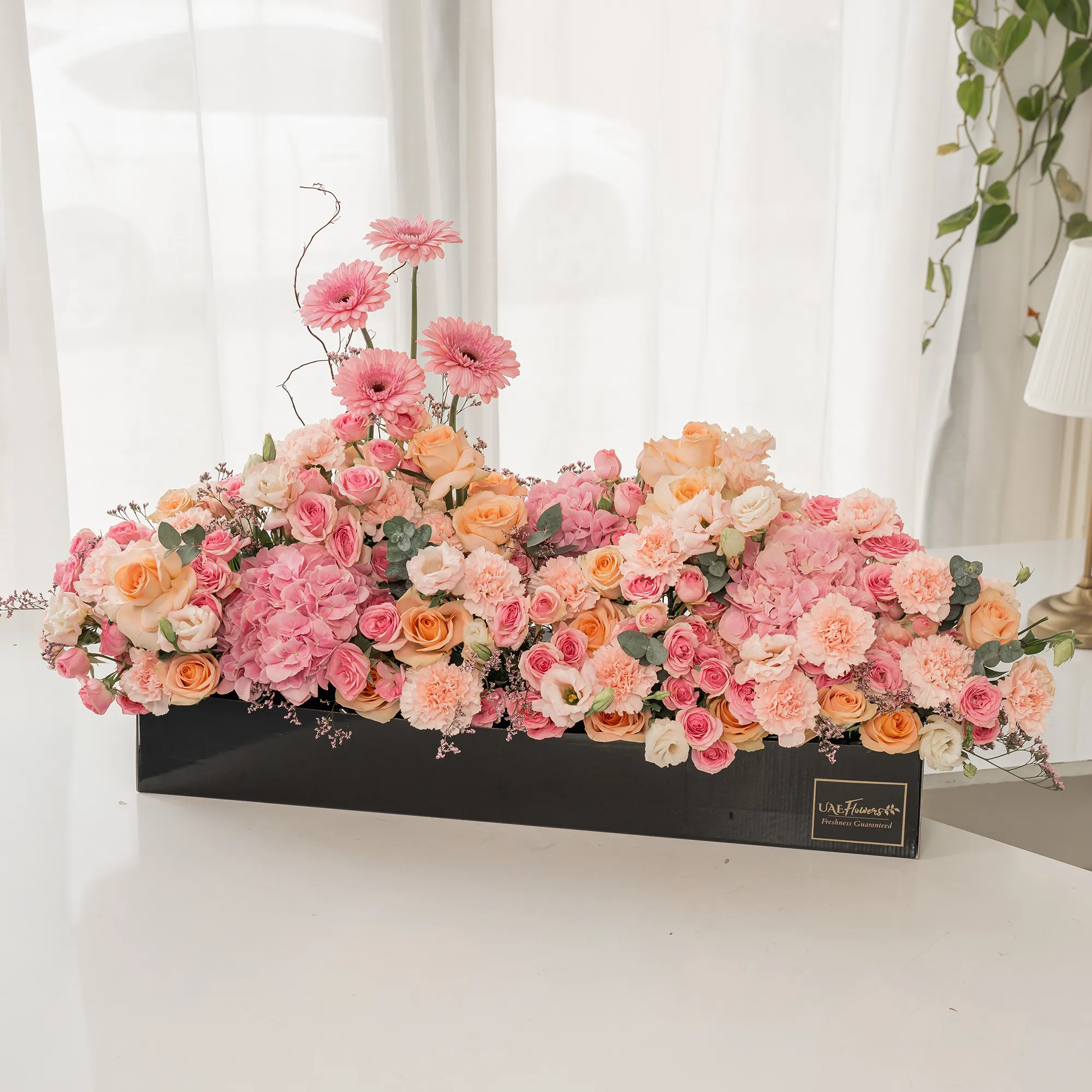Arrangement of 4 pink gerberas, 3 pink hydrangeas, 20 pink carnations, 20 pink spray roses and 30 peach roses with green fillers in a long black box.