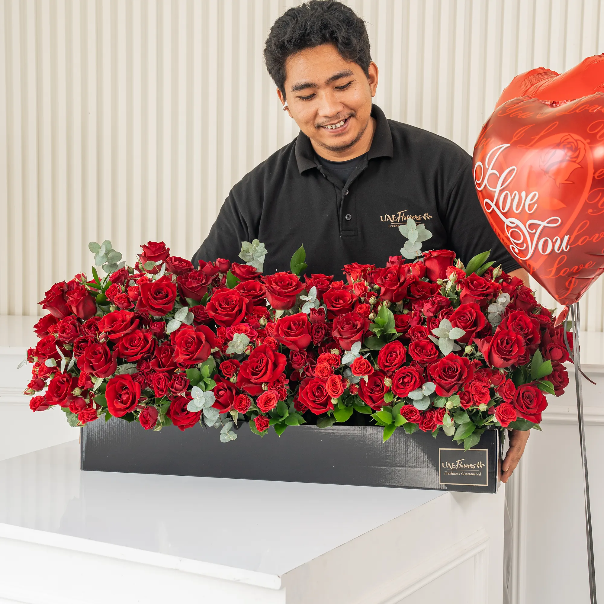 Arrangement of 50 red roses and 30 red spray roses with green fillers in a long black box.