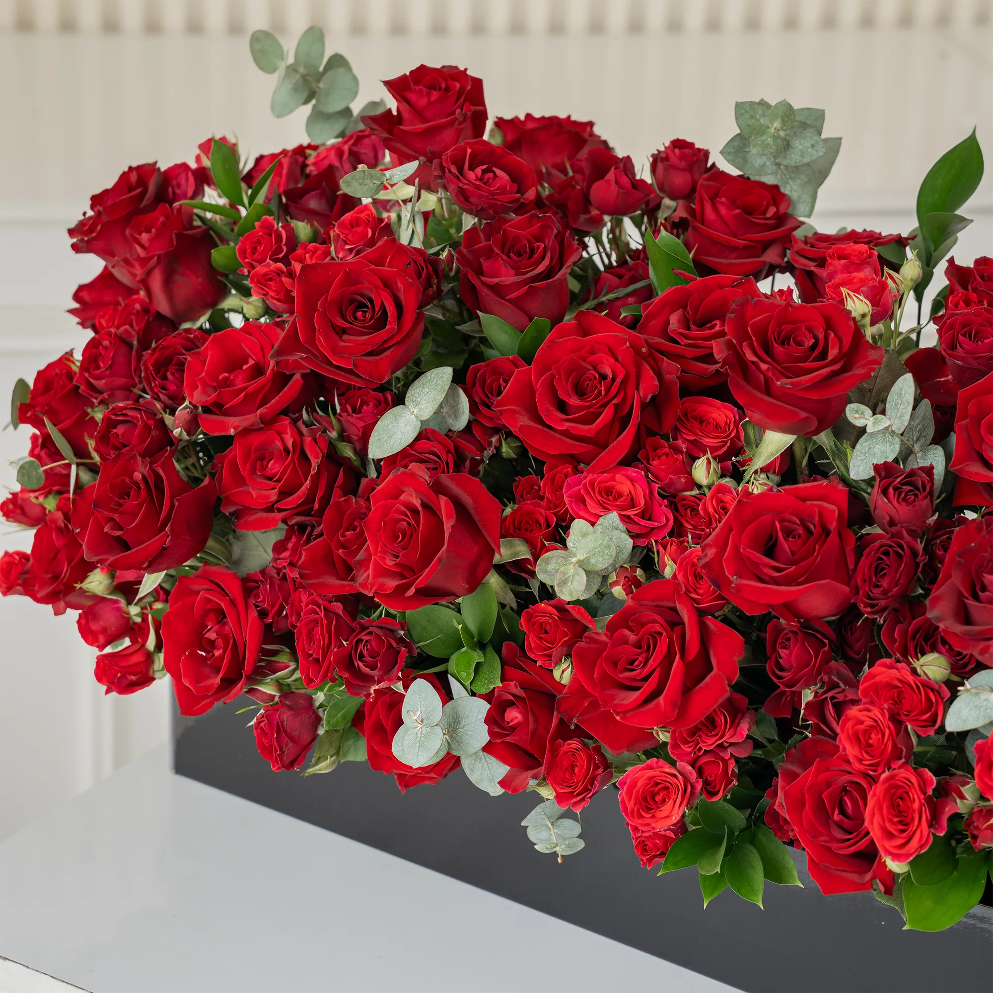 Arrangement of 50 red roses and 30 red spray roses with green fillers in a long black box.