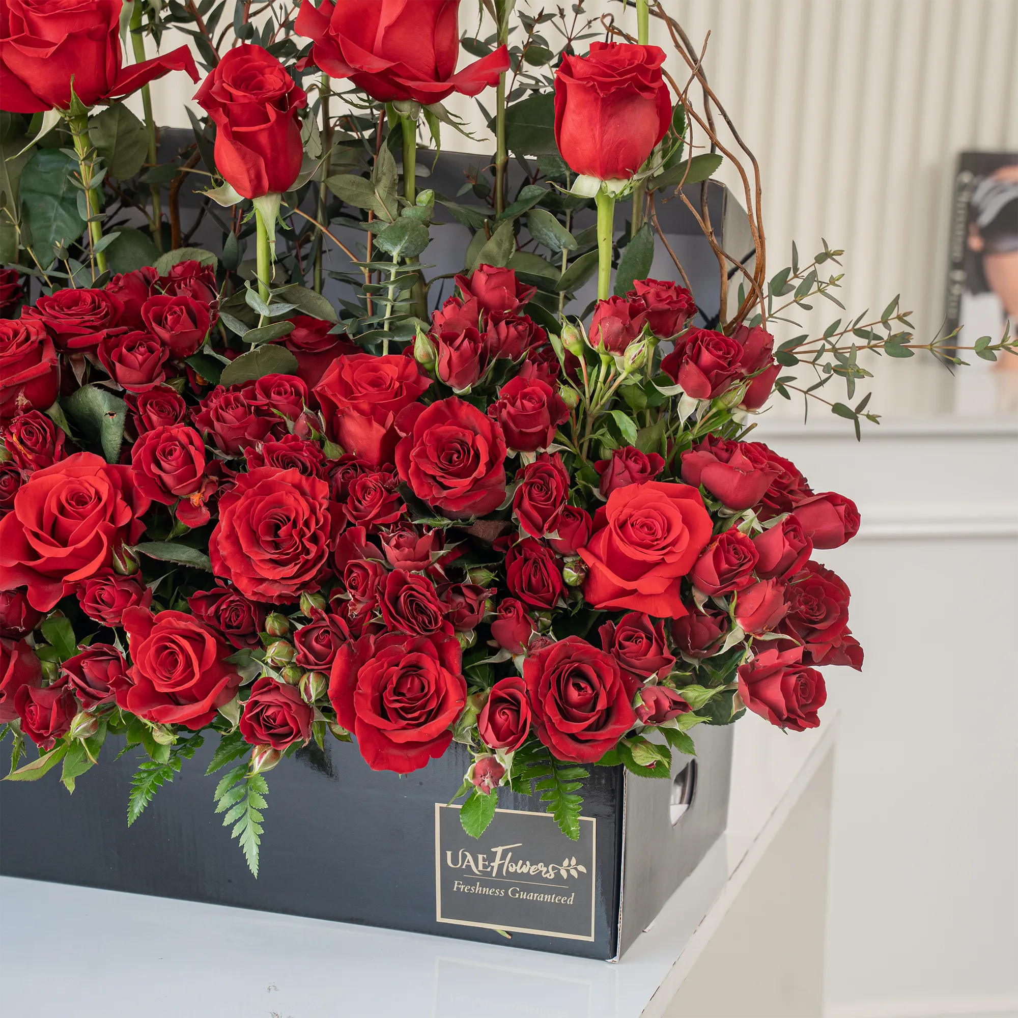 Arrangement of 50 red roses and 20 red spray roses with green fillers in a long black box.