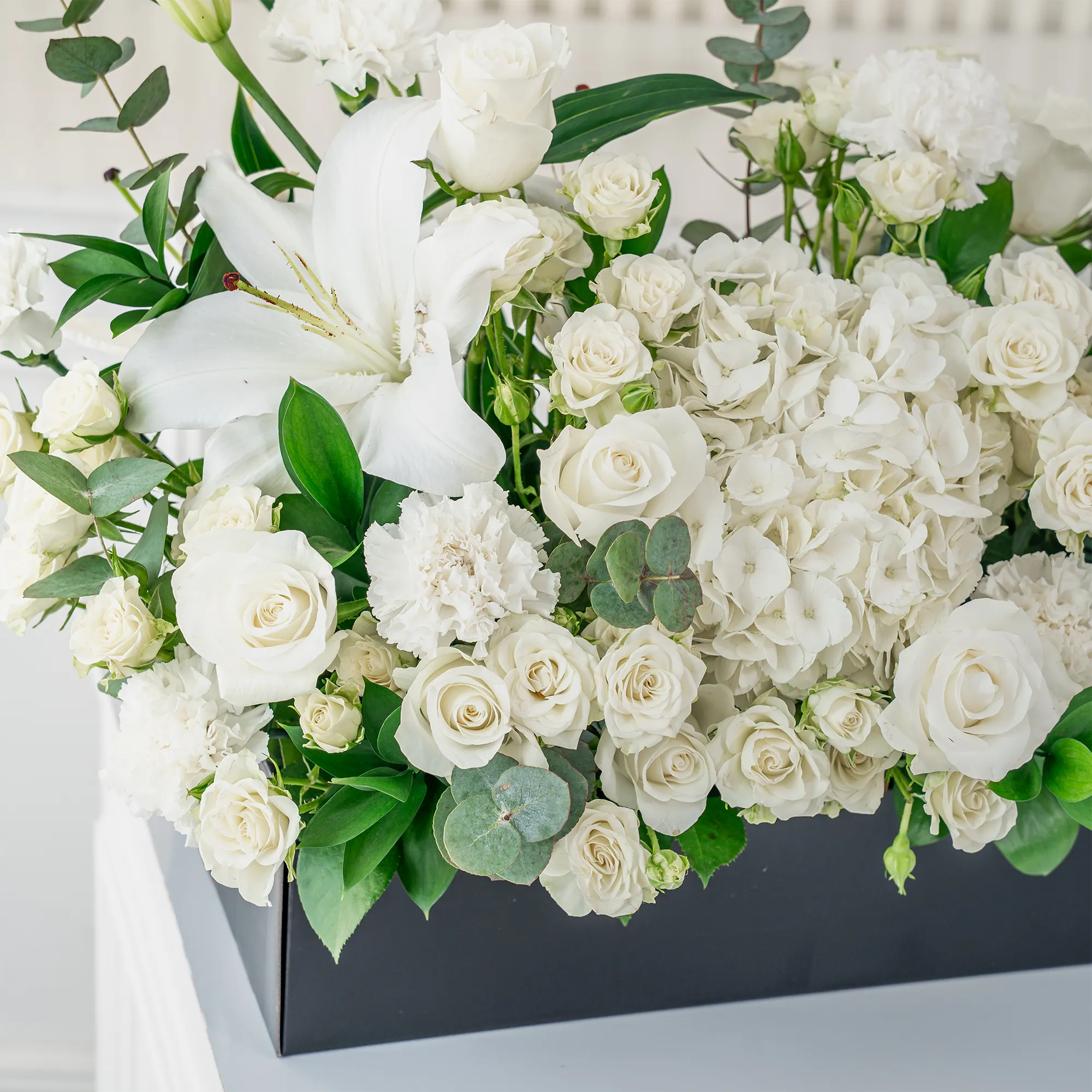 Arrangement of 5 white lilies, 20 white roses, 20 white spray roses, 10 white carnations and 2 white hydrangeas with green fillers in a long black box.