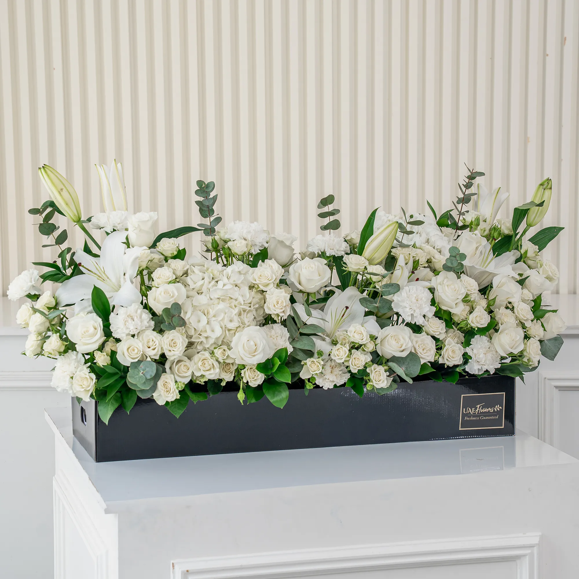 Arrangement of 5 white lilies, 20 white roses, 20 white spray roses, 10 white carnations and 2 white hydrangeas with green fillers in a long black box.
