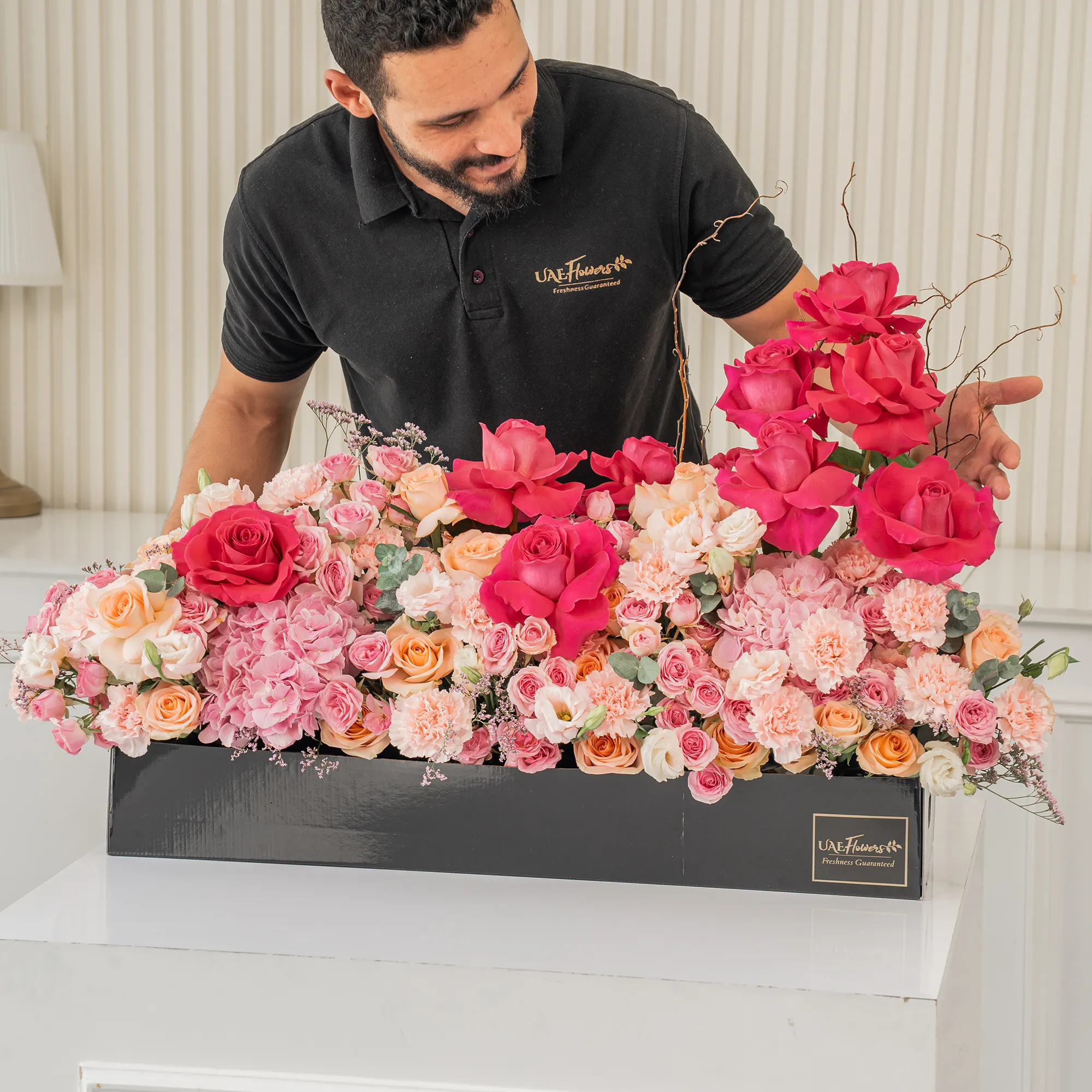 Arrangement of 10 fuchsia roses, 20 pink carnations, 20 pink spray roses, 20 peach roses and 2 pink hydrangeas with green fillers in a long black box.
