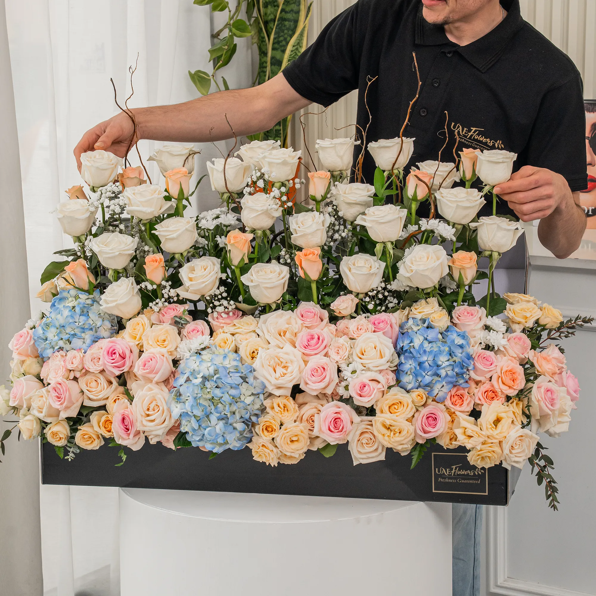 Arrangement of 30 White roses, 20 pink roses, 20 peach roses, 3 blue hydrangeas and 10 peach spray roses with green fillers in a long black box.