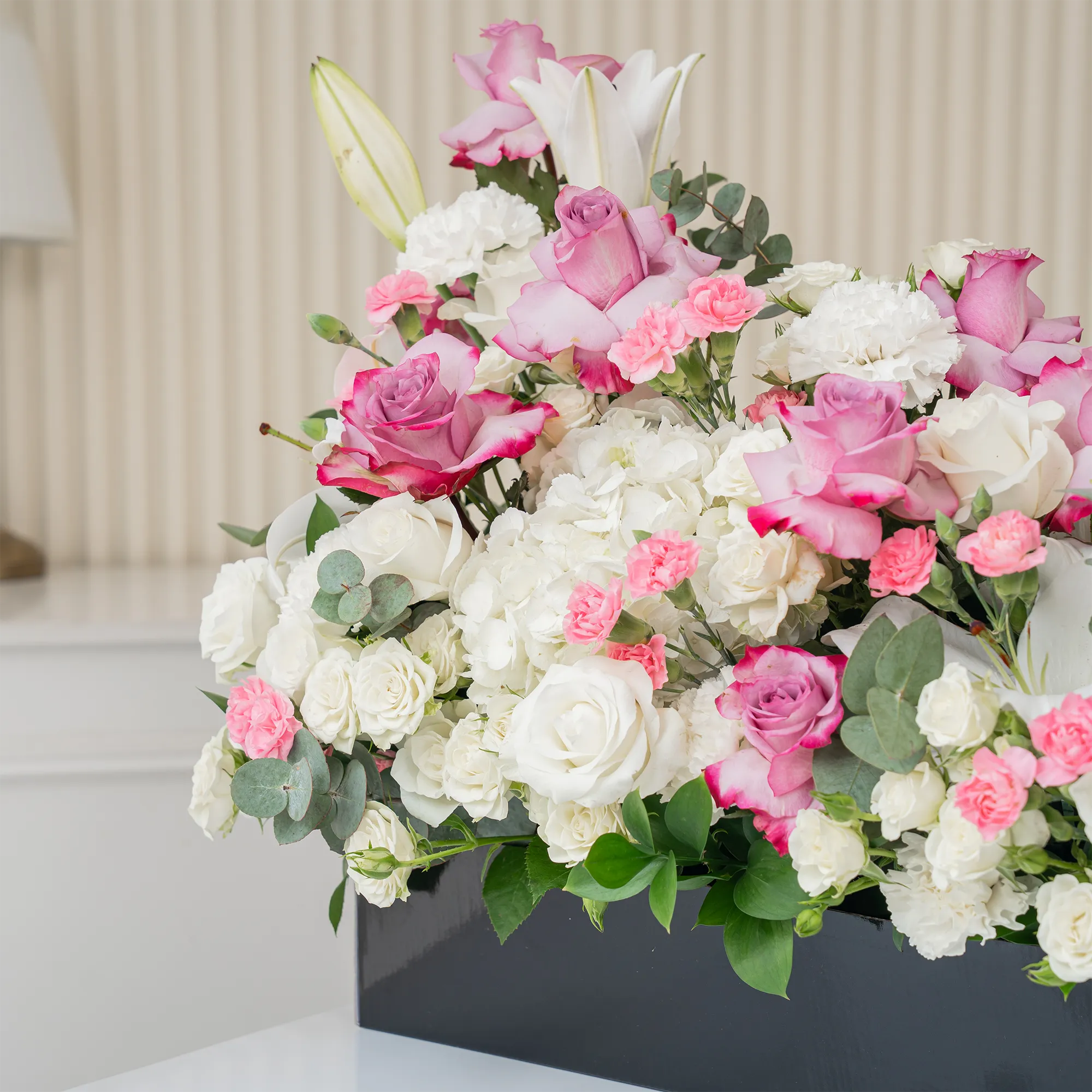 Arrangement of 5 white lilies, 15 purple roses, 15 white roses, 10 white carnations, 10 pink carnations, 10 white spray roses and 2 white hydrangeas with green fillers in a long black box.