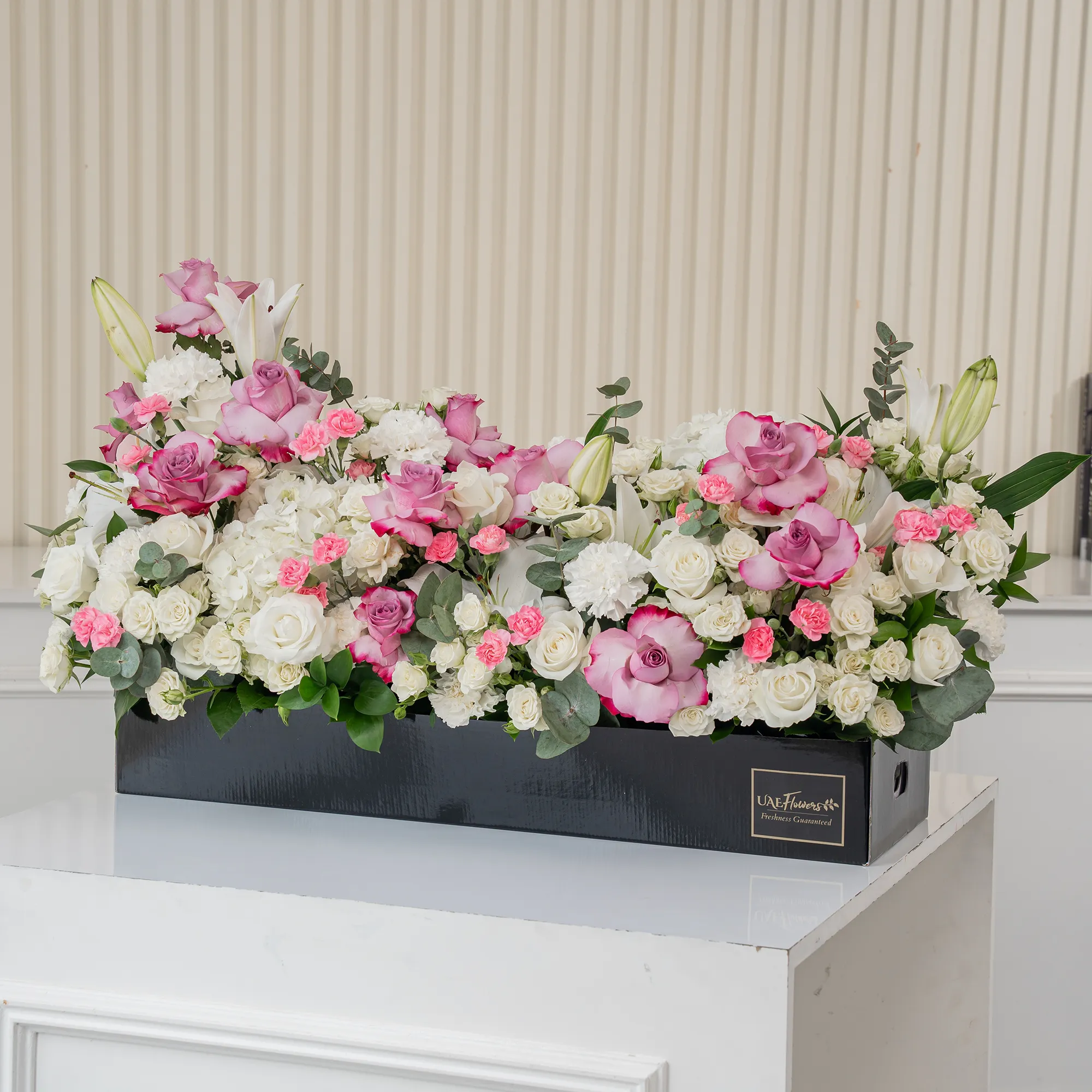 Arrangement of 5 white lilies, 15 purple roses, 15 white roses, 10 white carnations, 10 pink carnations, 10 white spray roses and 2 white hydrangeas with green fillers in a long black box.