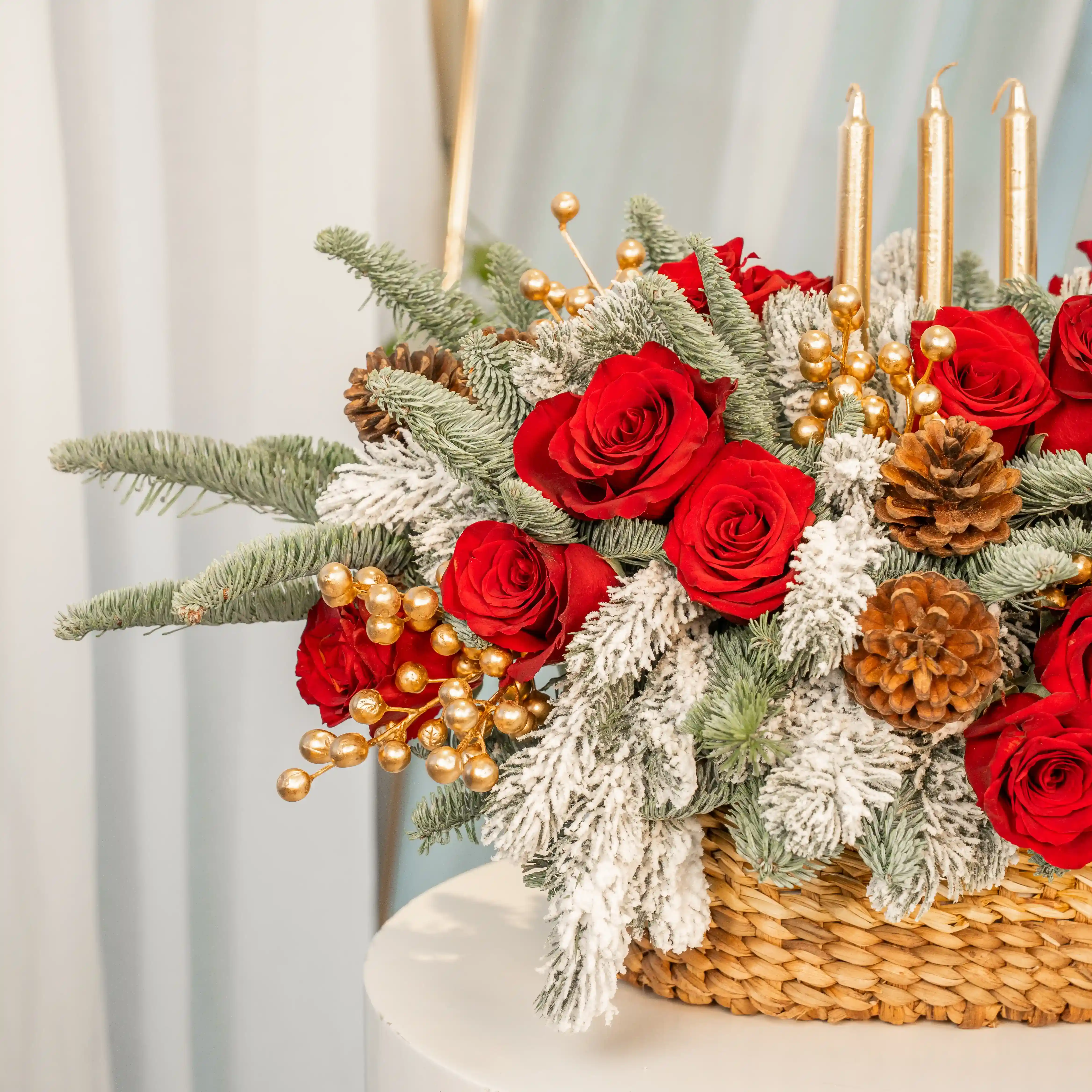 Christmas centerpiece with red roses, snowy pine, gold candles, pinecones in a woven basket