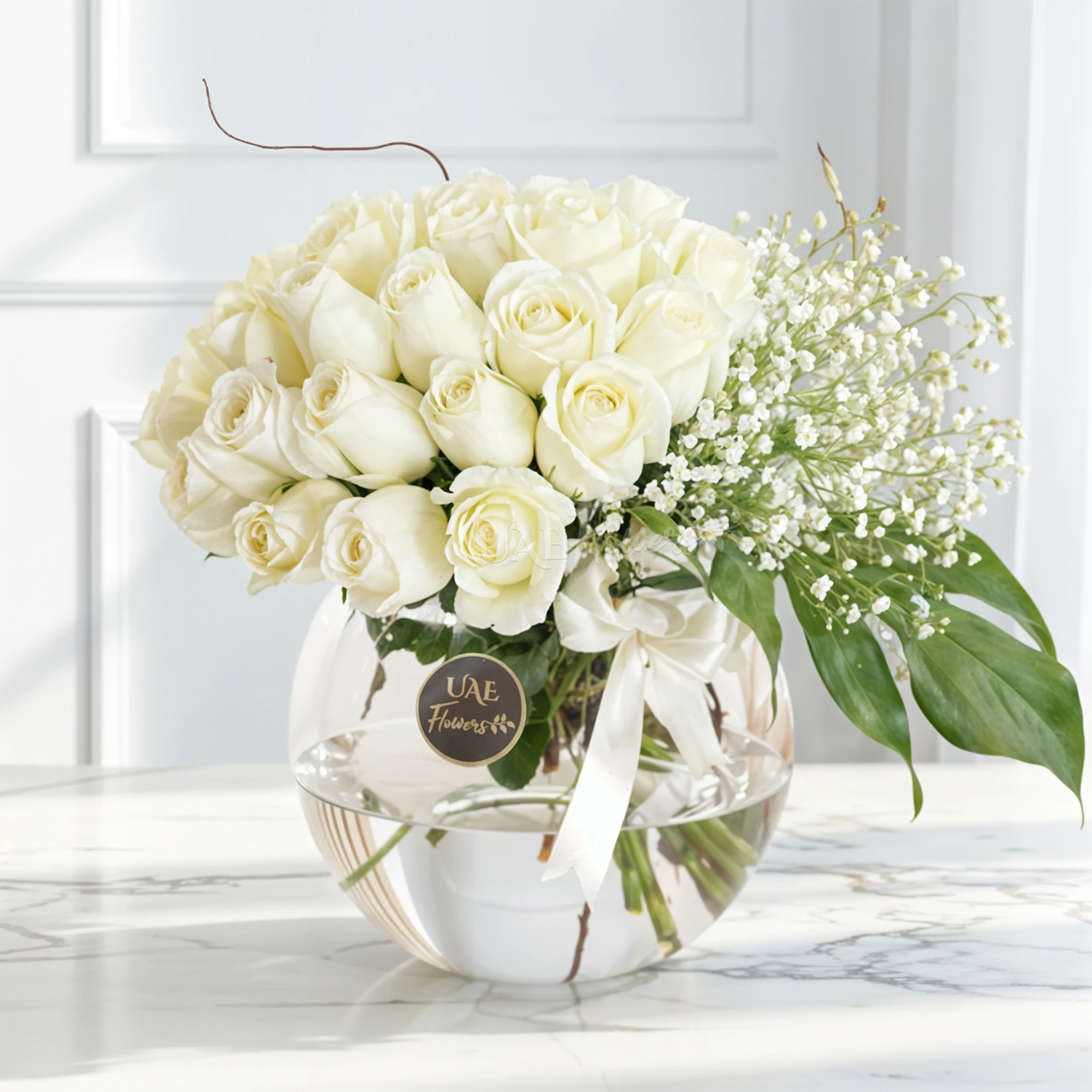 Round glass vase with 20 white roses, baby's breath, and green fillers, elegantly arranged.
