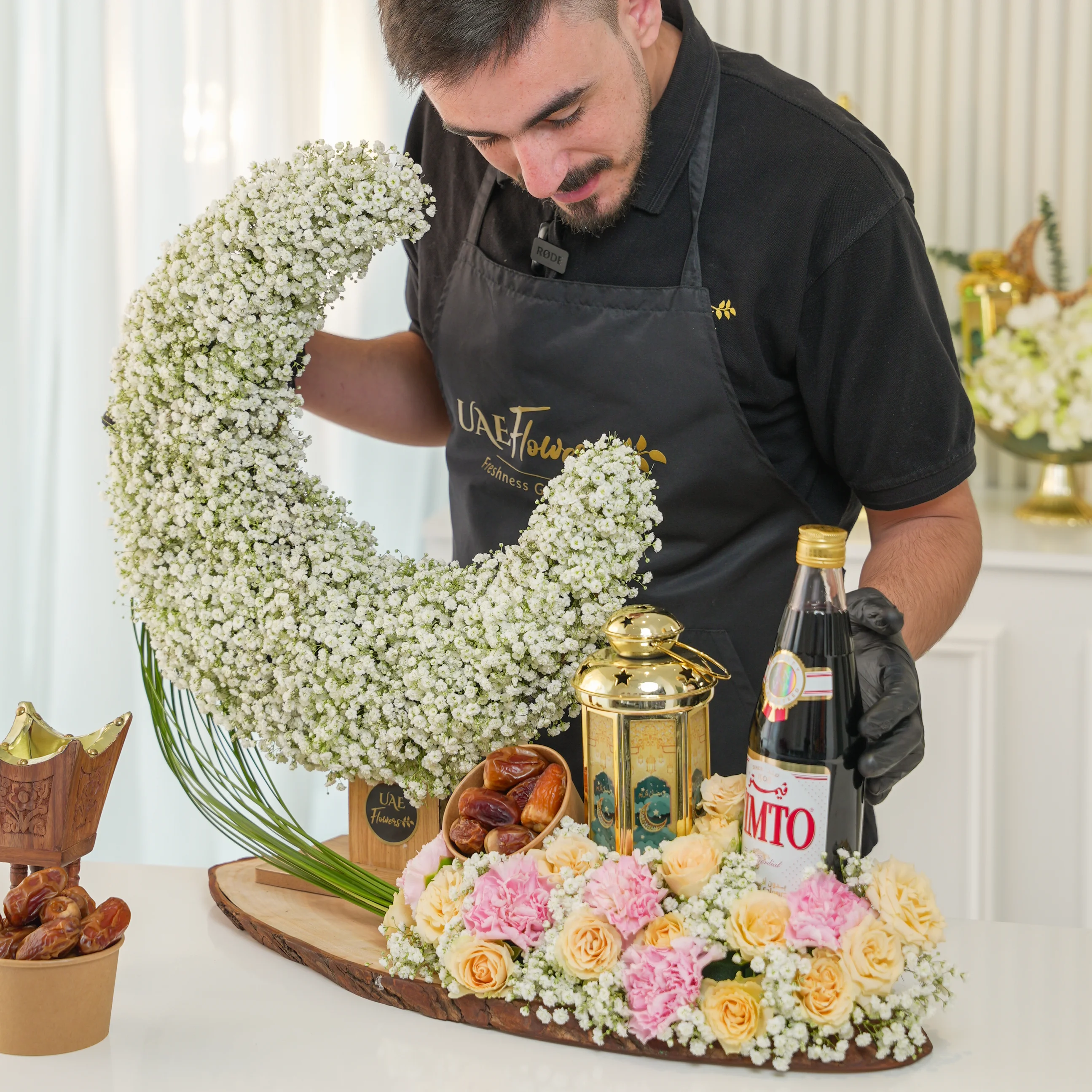 baby’s breath, yellow spray roses, pink carnations, dates, gold lantern, Vimto drink, and wooden base.