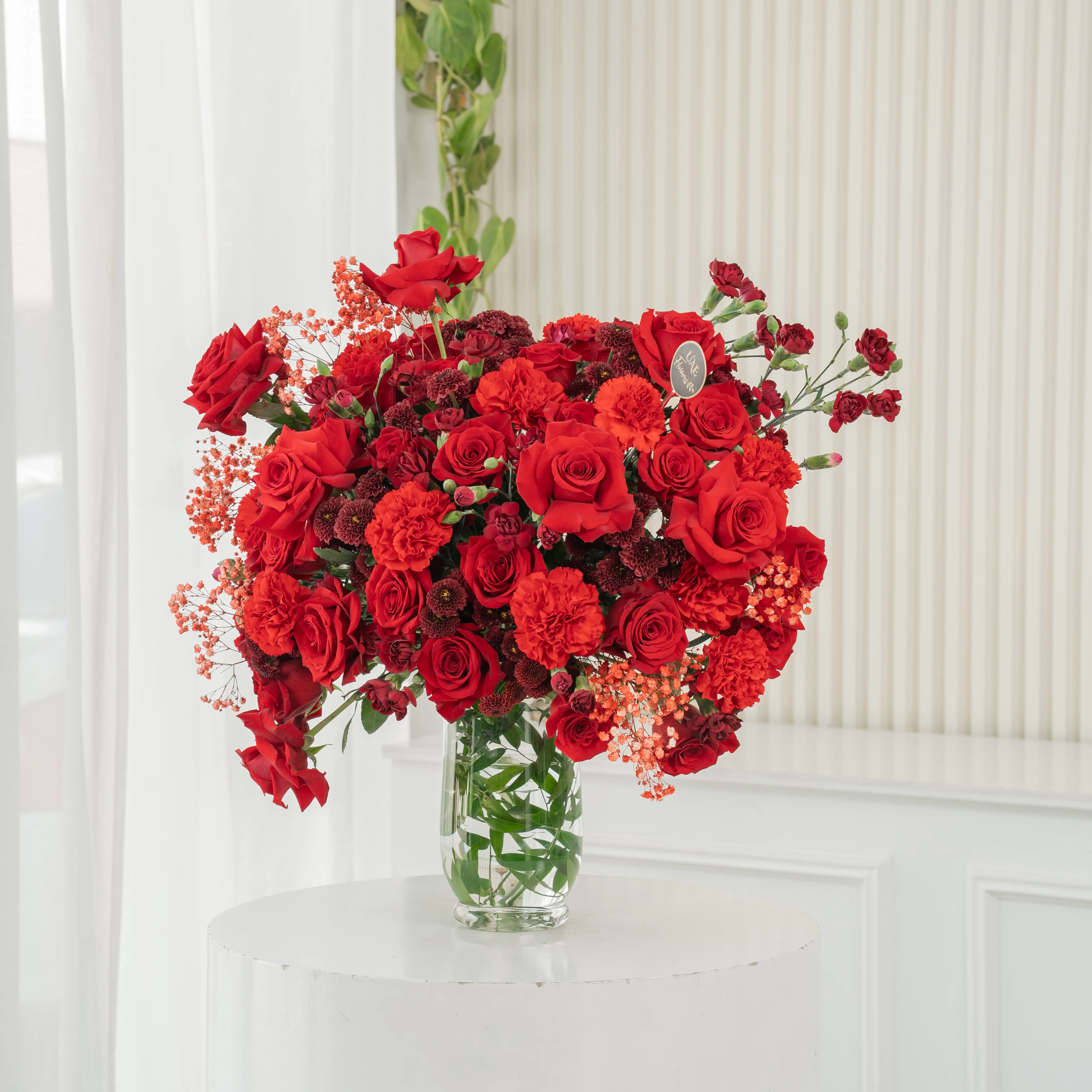 One-side arrangement of 20 red roses, 7 red chrysanthemums, 8 red carnations and 4 red spray carnations with green fillers in a glass vase.