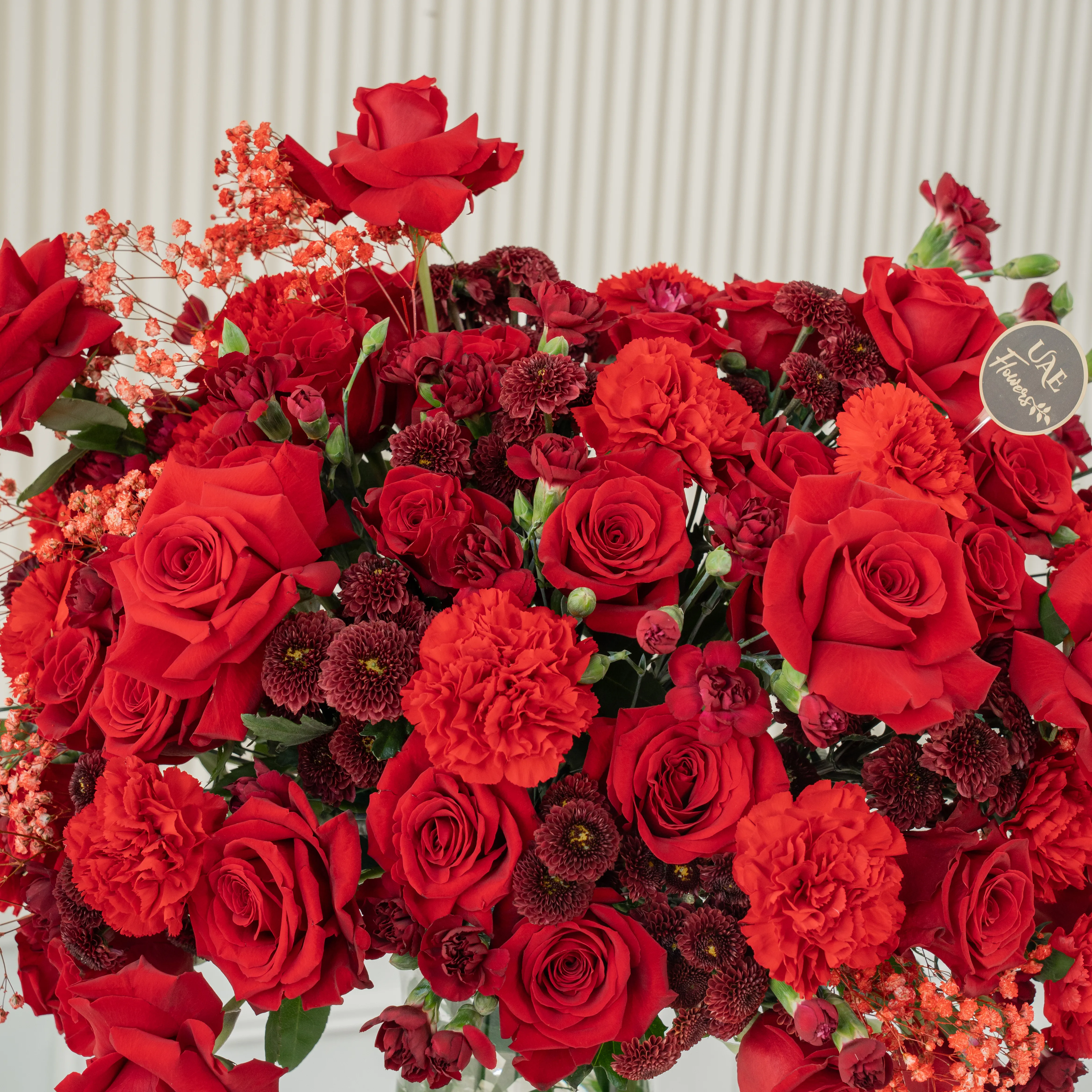 One-side arrangement of 20 red roses, 7 red chrysanthemums, 8 red carnations and 4 red spray carnations with green fillers in a glass vase.