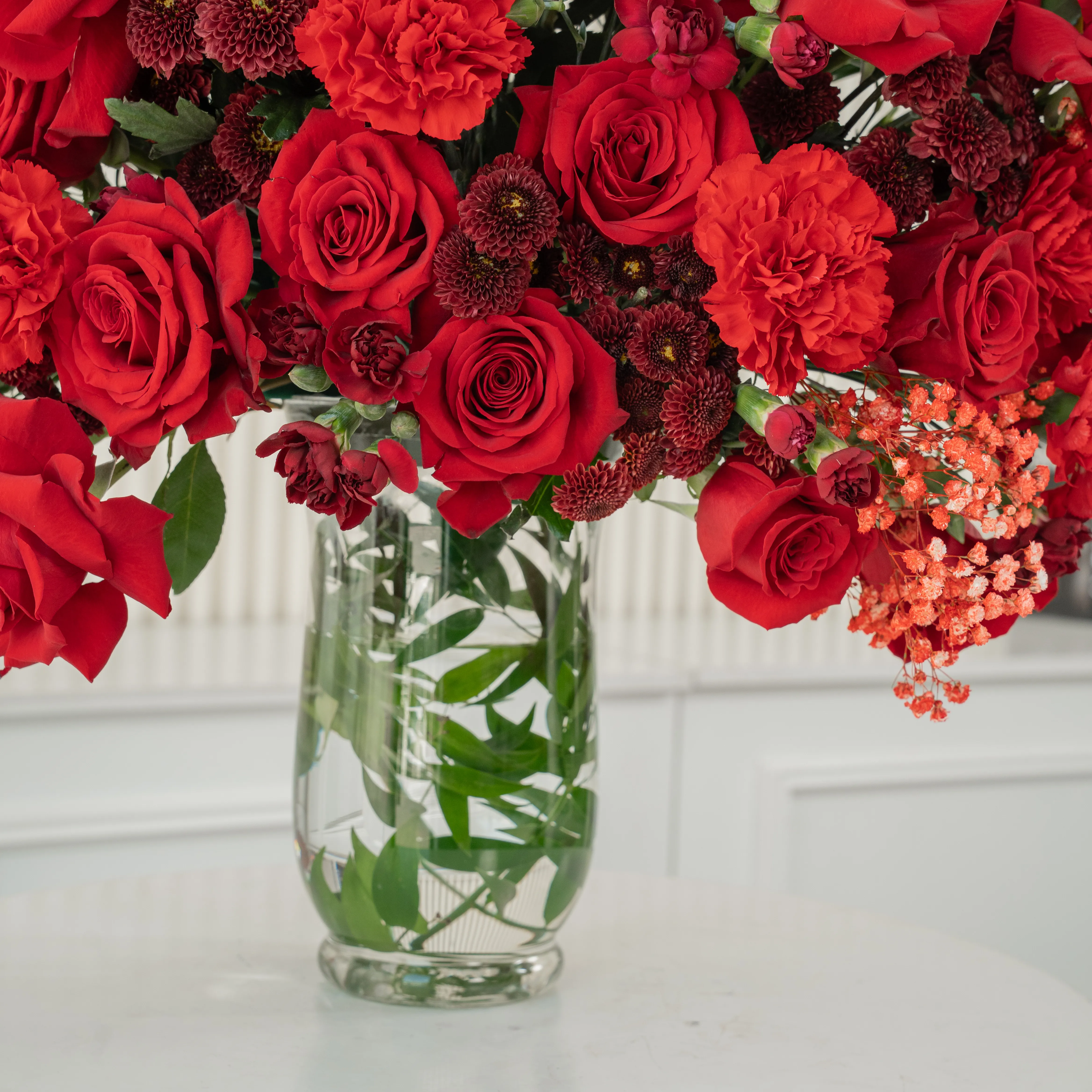 One-side arrangement of 20 red roses, 7 red chrysanthemums, 8 red carnations and 4 red spray carnations with green fillers in a glass vase.