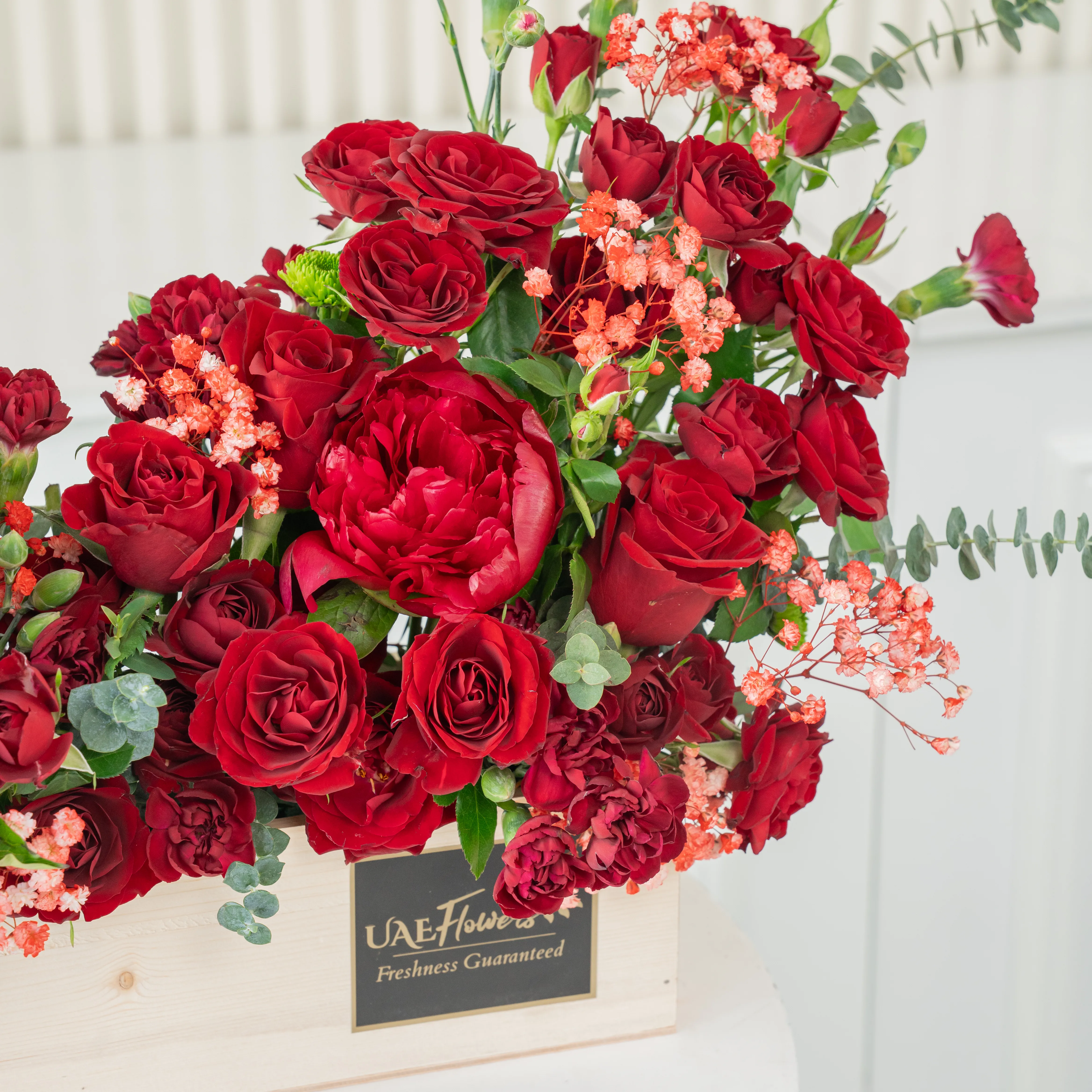 An arrangement of 10 red roses, 10 red spray roses and 3 red peonies with green fillers in a wooden tray.