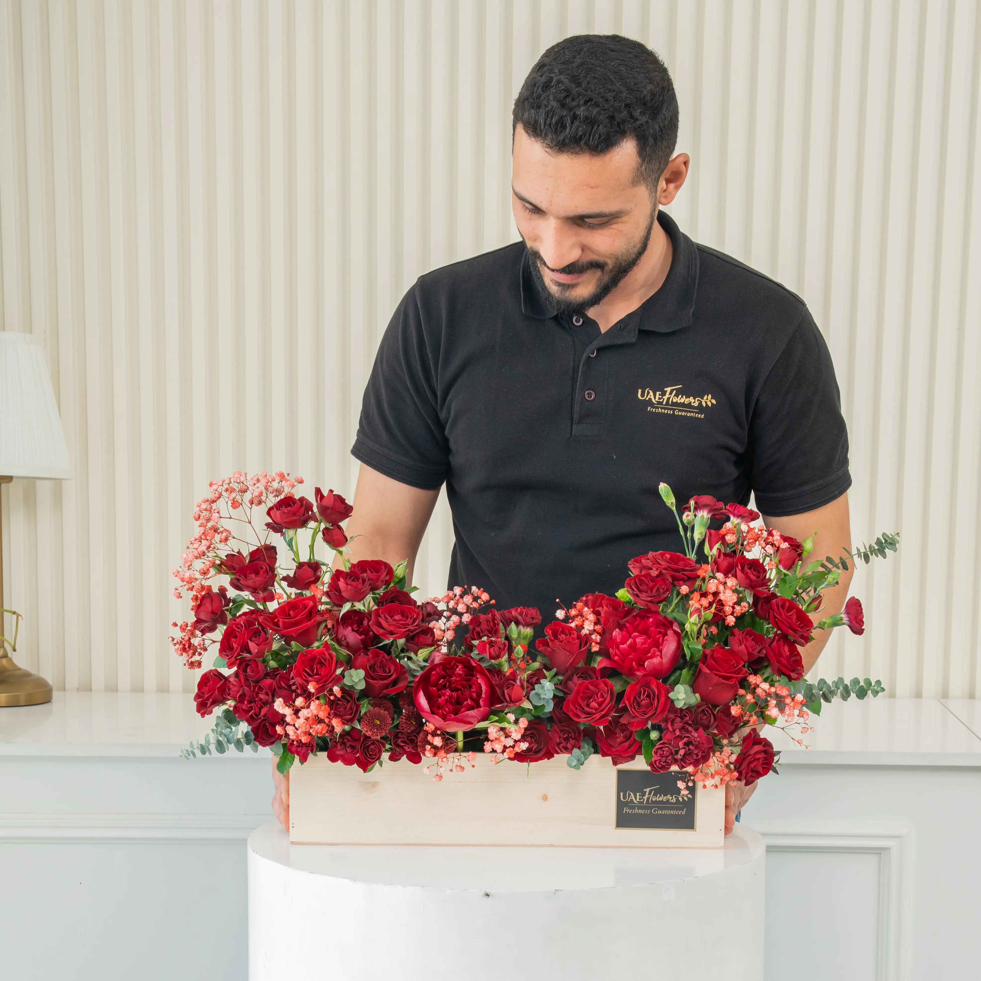 An arrangement of 10 red roses, 10 red spray roses and 3 red peonies with green fillers in a wooden tray.