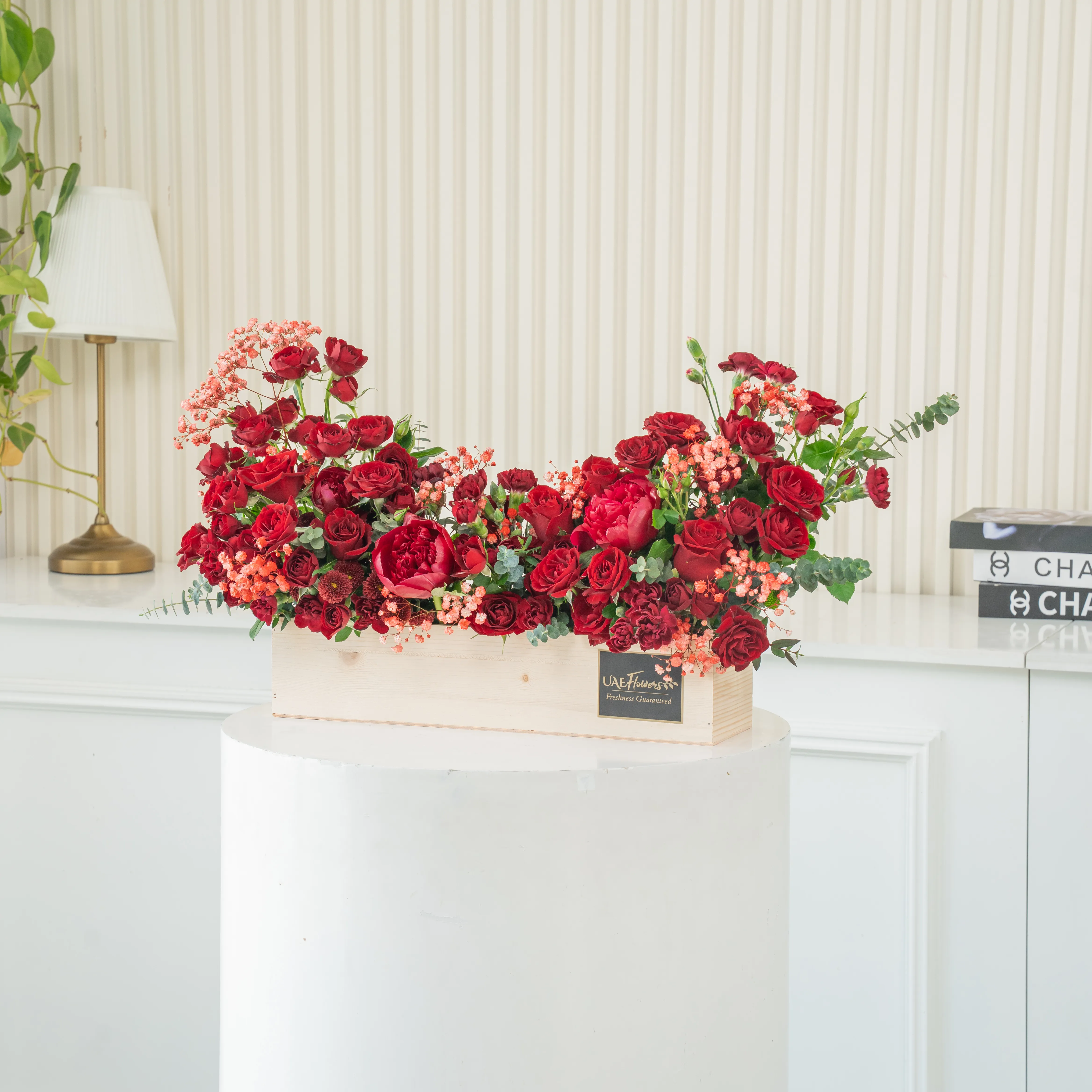 An arrangement of 10 red roses, 10 red spray roses and 3 red peonies with green fillers in a wooden tray.
