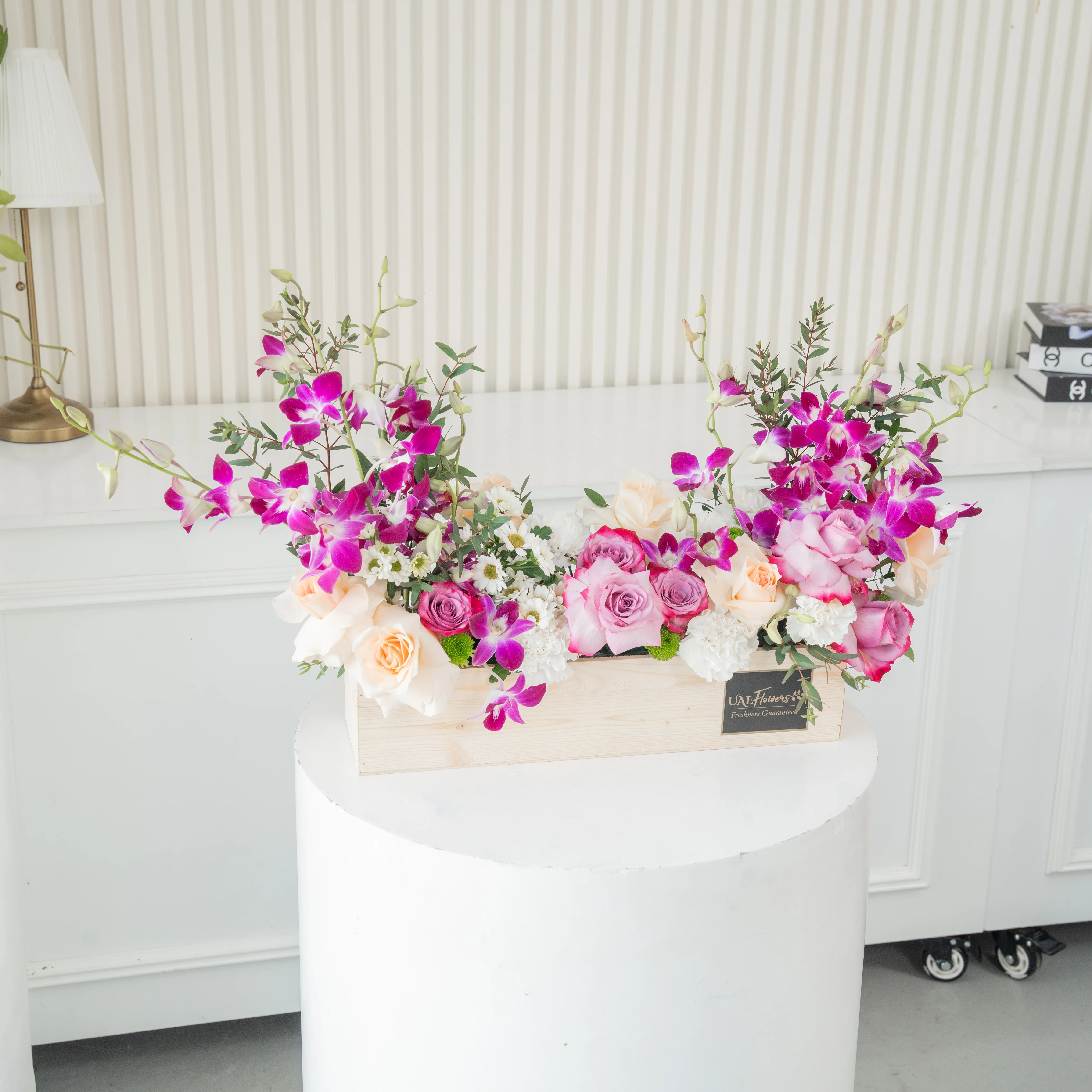 6 purple roses, 6 peach roses, 5 white carnations, 2 white chrysanthemums and 6 purple orchids in a wooden tray.