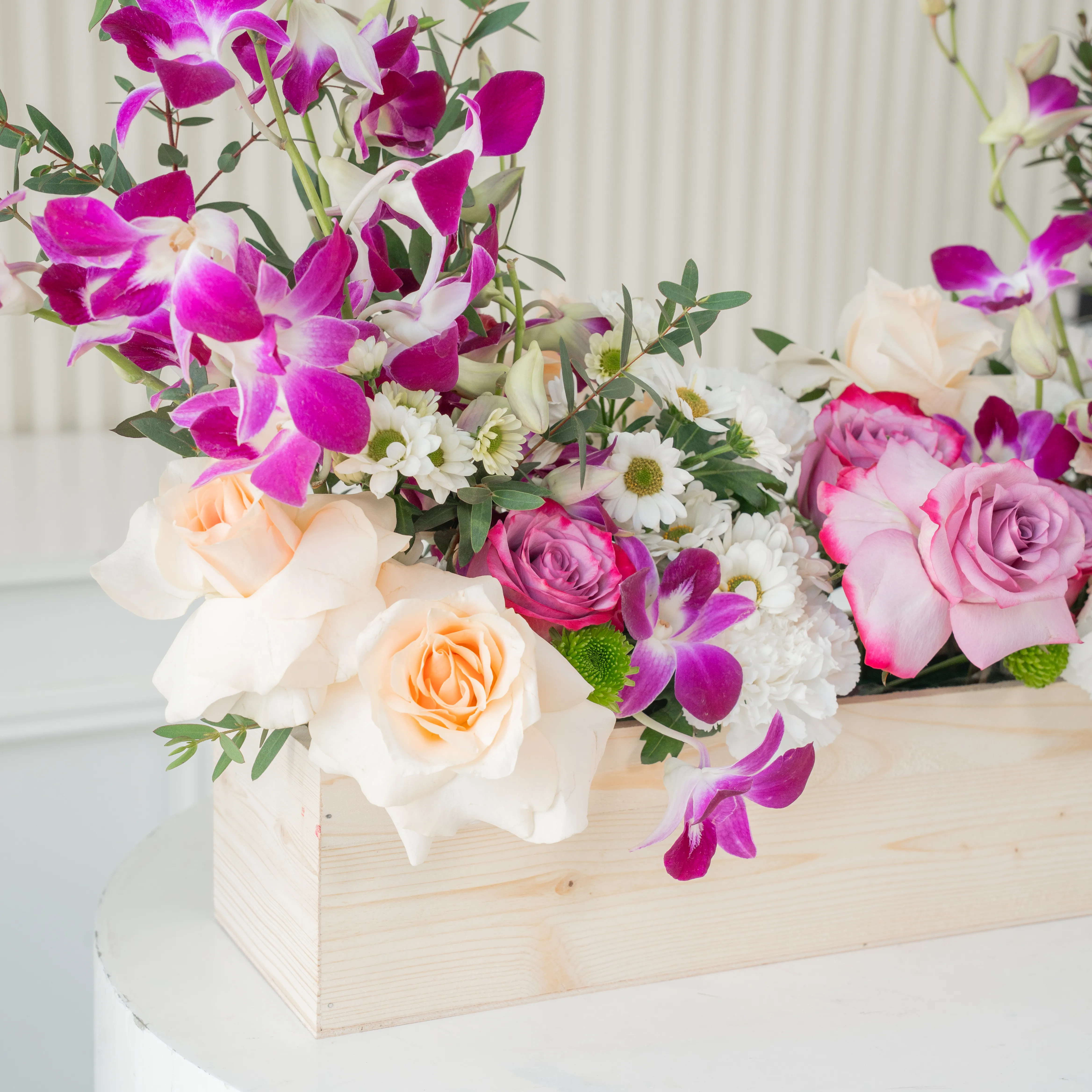 6 purple roses, 6 peach roses, 5 white carnations, 2 white chrysanthemums and 6 purple orchids in a wooden tray.