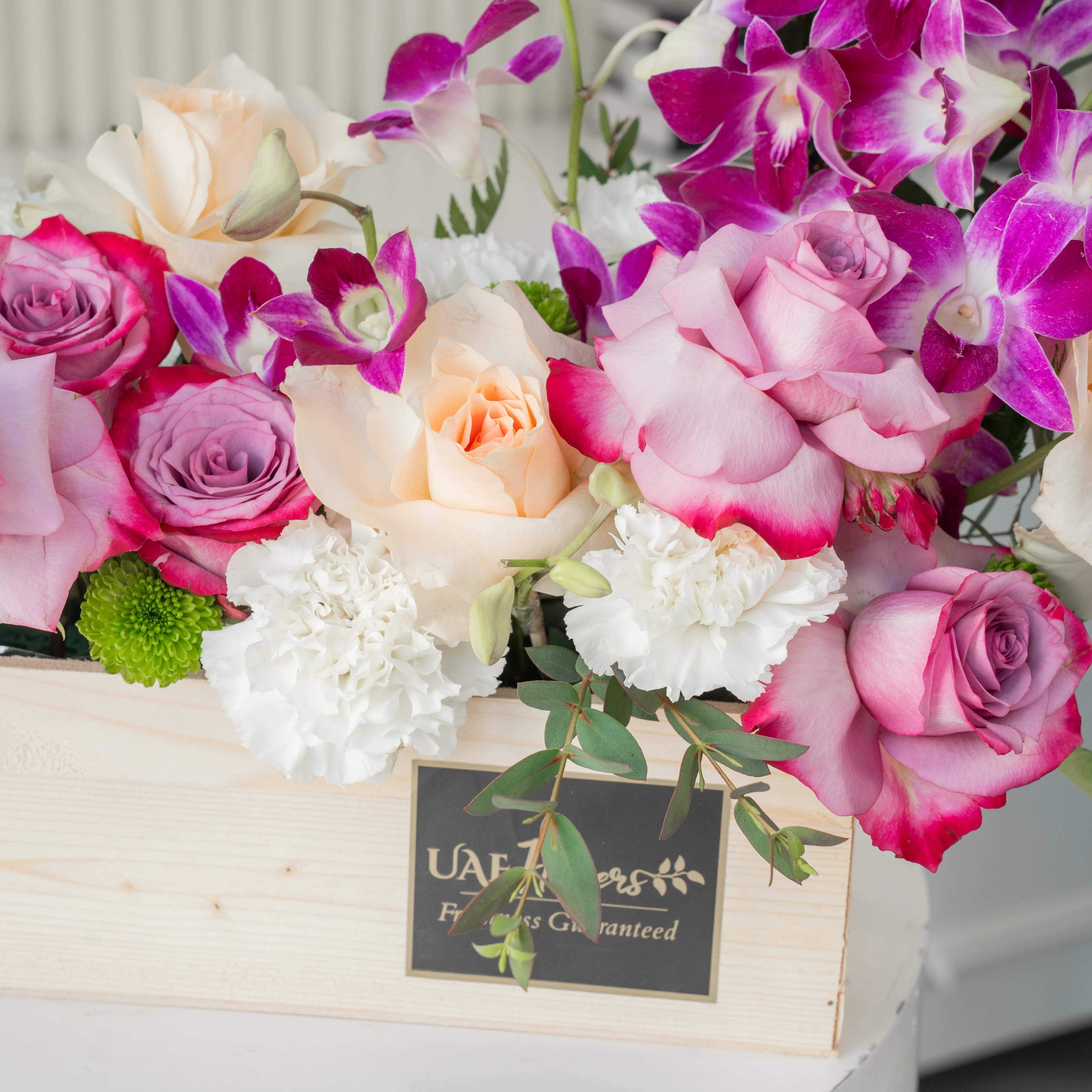 6 purple roses, 6 peach roses, 5 white carnations, 2 white chrysanthemums and 6 purple orchids in a wooden tray.