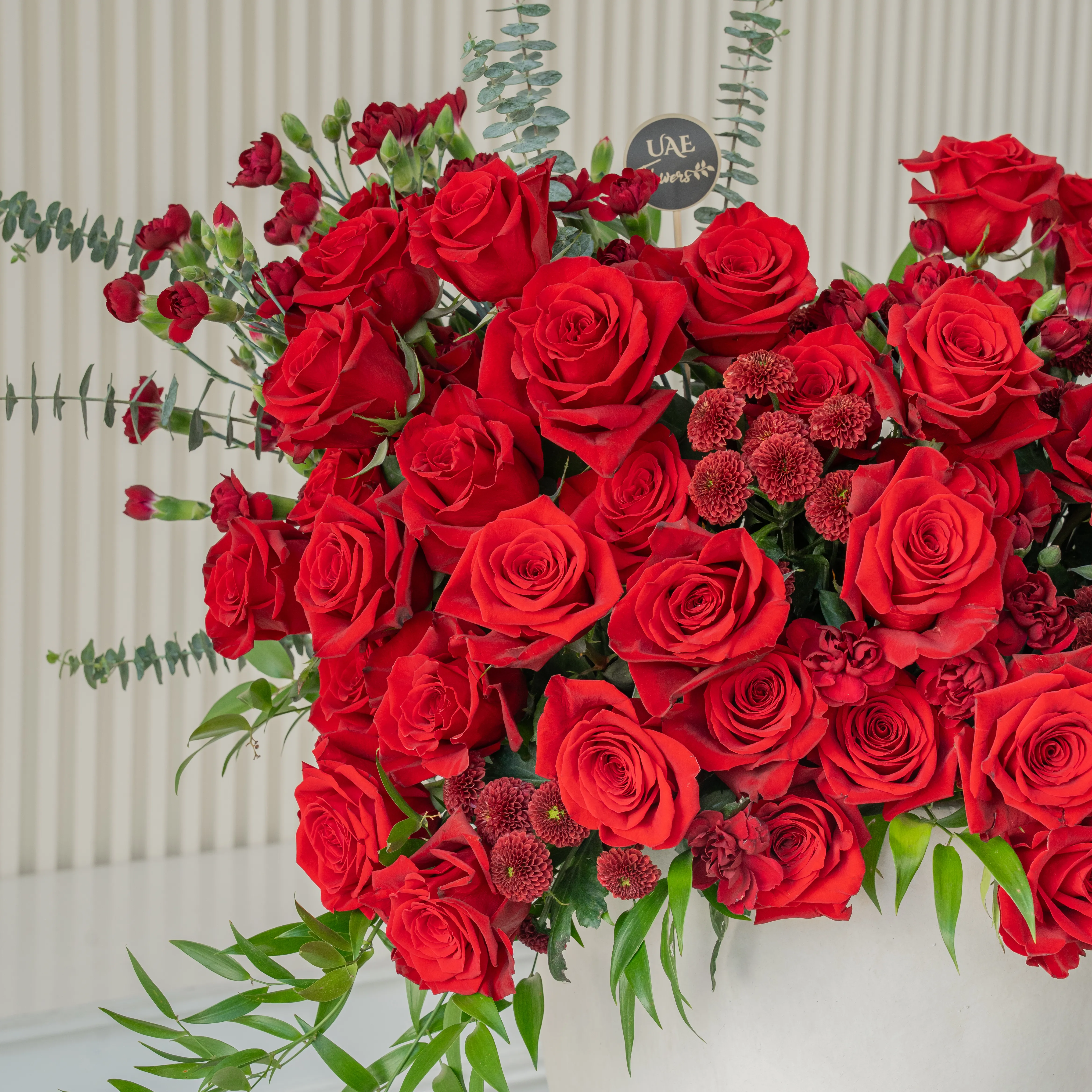 One-side arrangement of 50 red roses, 5 red spray carnations, 5 maroon button chrysanthemums & 4 Black Annie Orchids with green fillers in a ceramic vase.