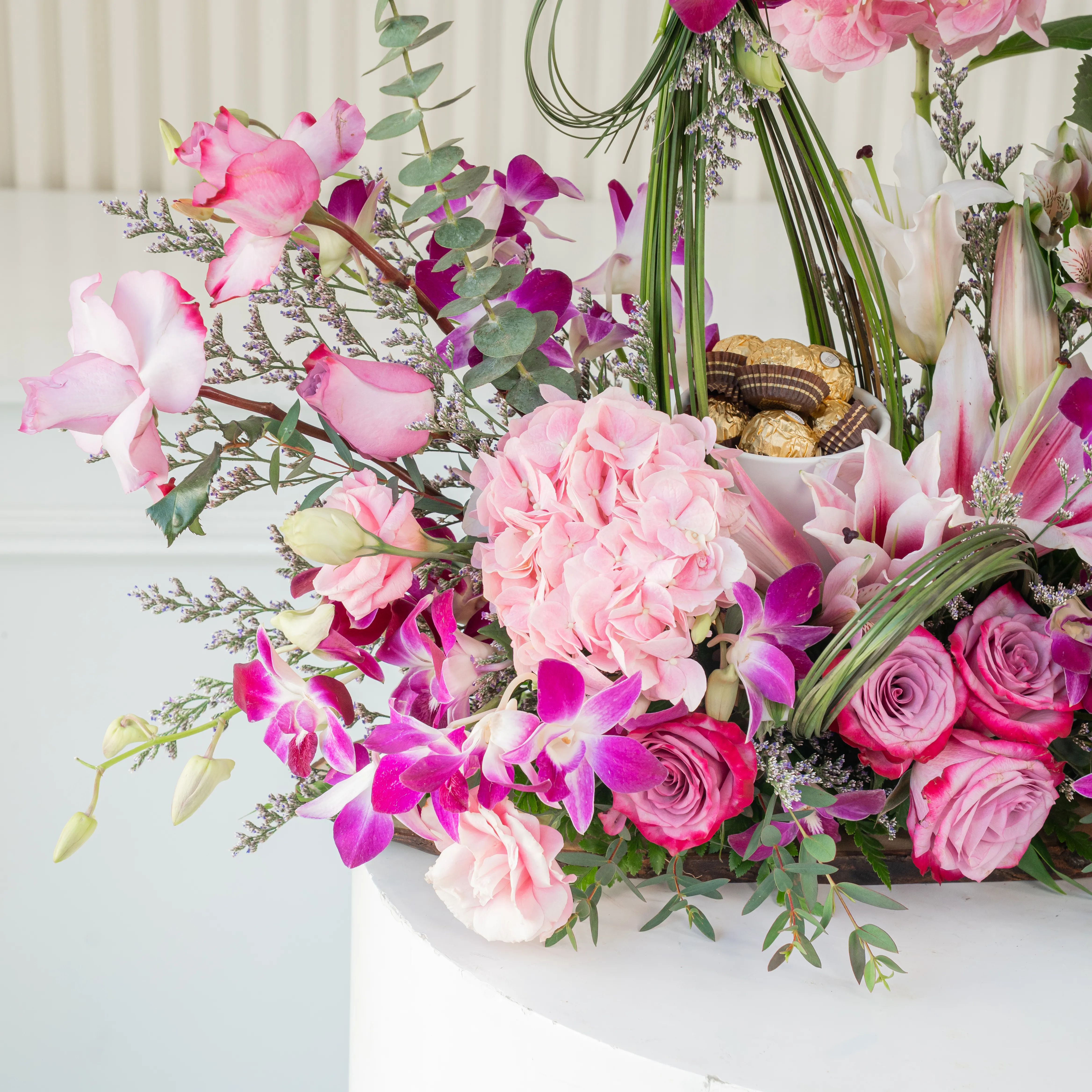 An arrangement of 12 purple roses, 15 purple orchids, 1 pink lily, 2 pink alstromeria, 5 pink lisianthus, 3 pink hydrangeas, bunch of steel grass and 8pcs Ferrero rocher chocolate with green fillers on a wooden log.