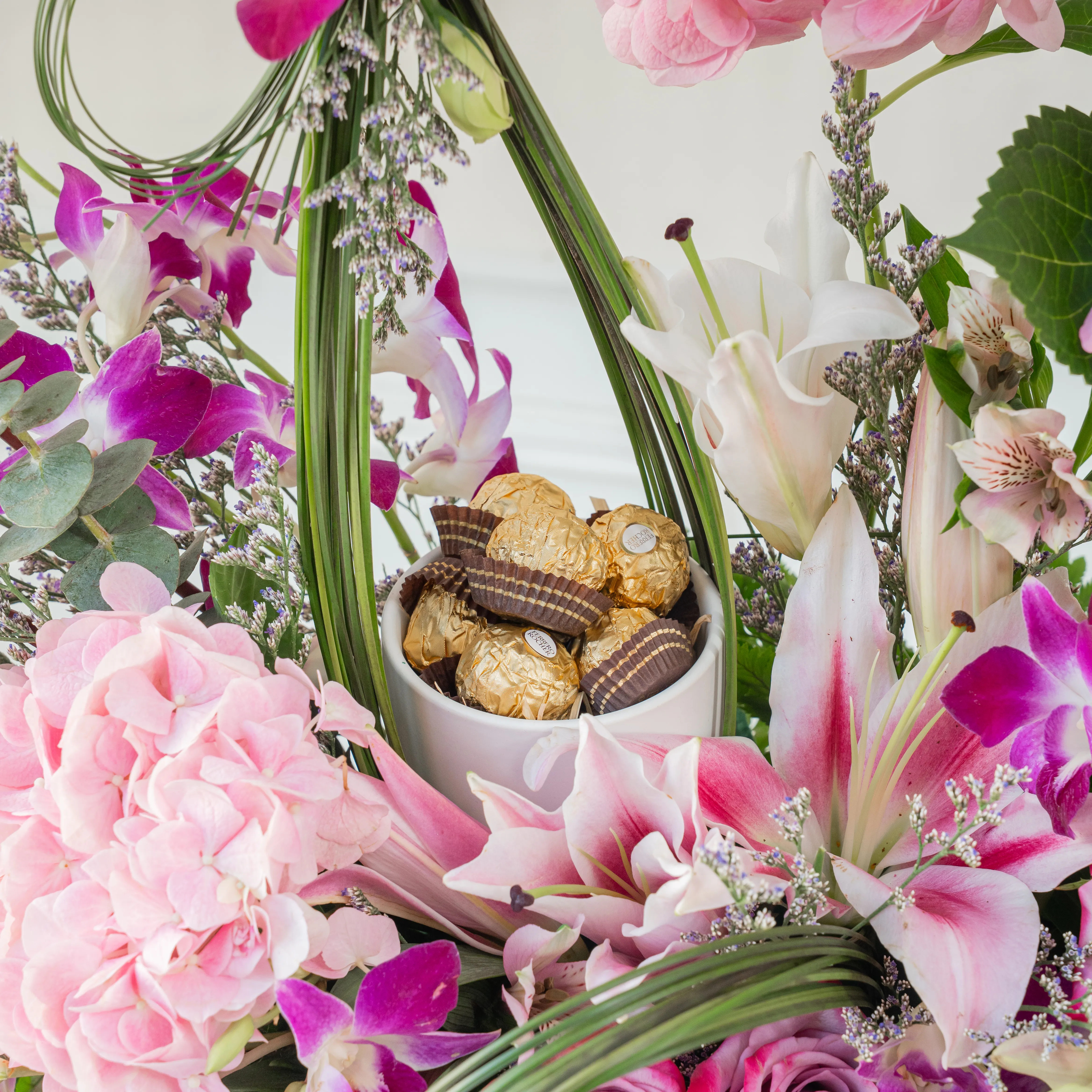 An arrangement of 12 purple roses, 15 purple orchids, 1 pink lily, 2 pink alstromeria, 5 pink lisianthus, 3 pink hydrangeas, bunch of steel grass and 8pcs Ferrero rocher chocolate with green fillers on a wooden log.