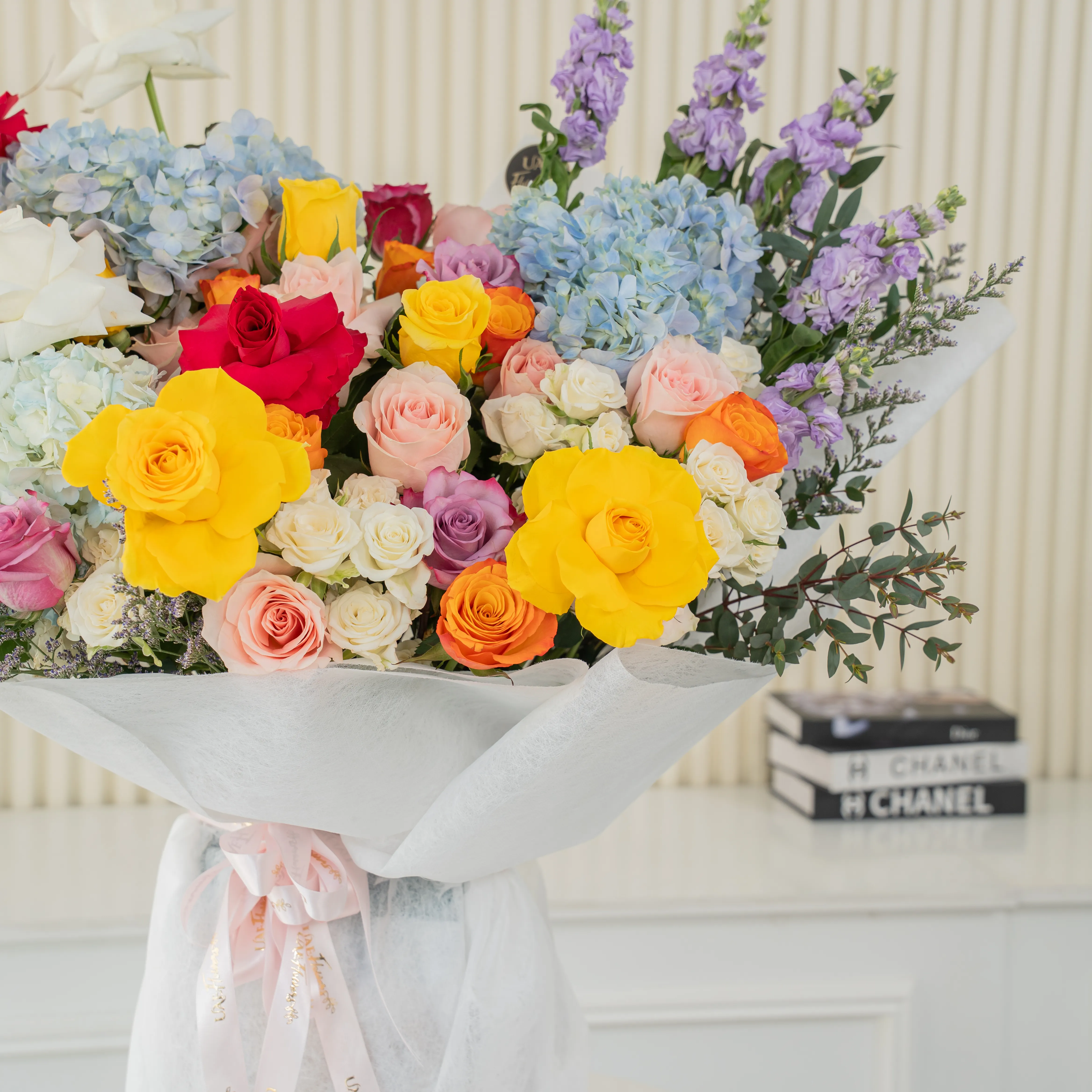 Bouquet of 25 mix roses, 6 white spray roses, 3 blue hydrangeas and 5 purple matthiola with green fillers in white paper packing tied with pink ribbon.