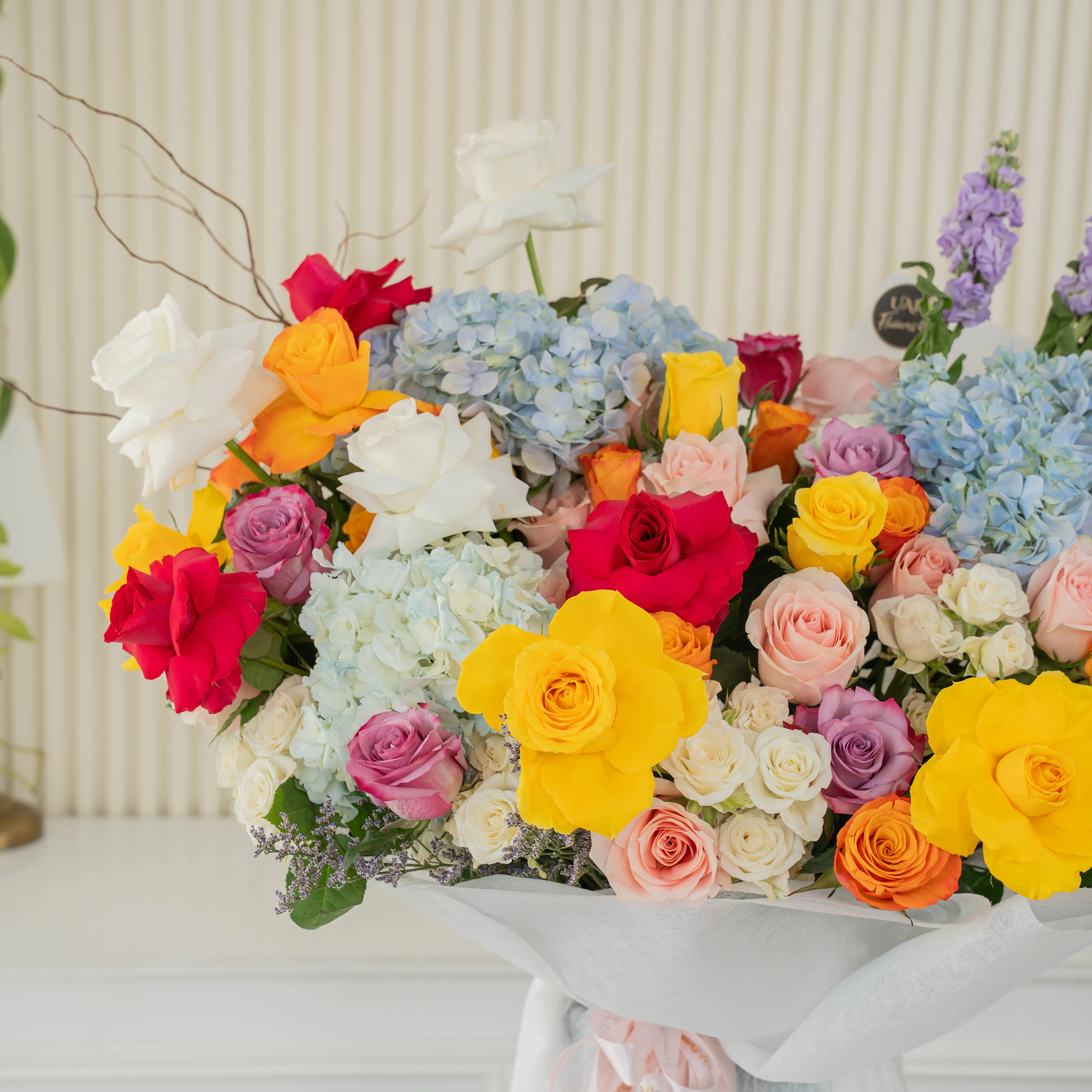 Bouquet of 25 mix roses, 6 white spray roses, 3 blue hydrangeas and 5 purple matthiola with green fillers in white paper packing tied with pink ribbon.