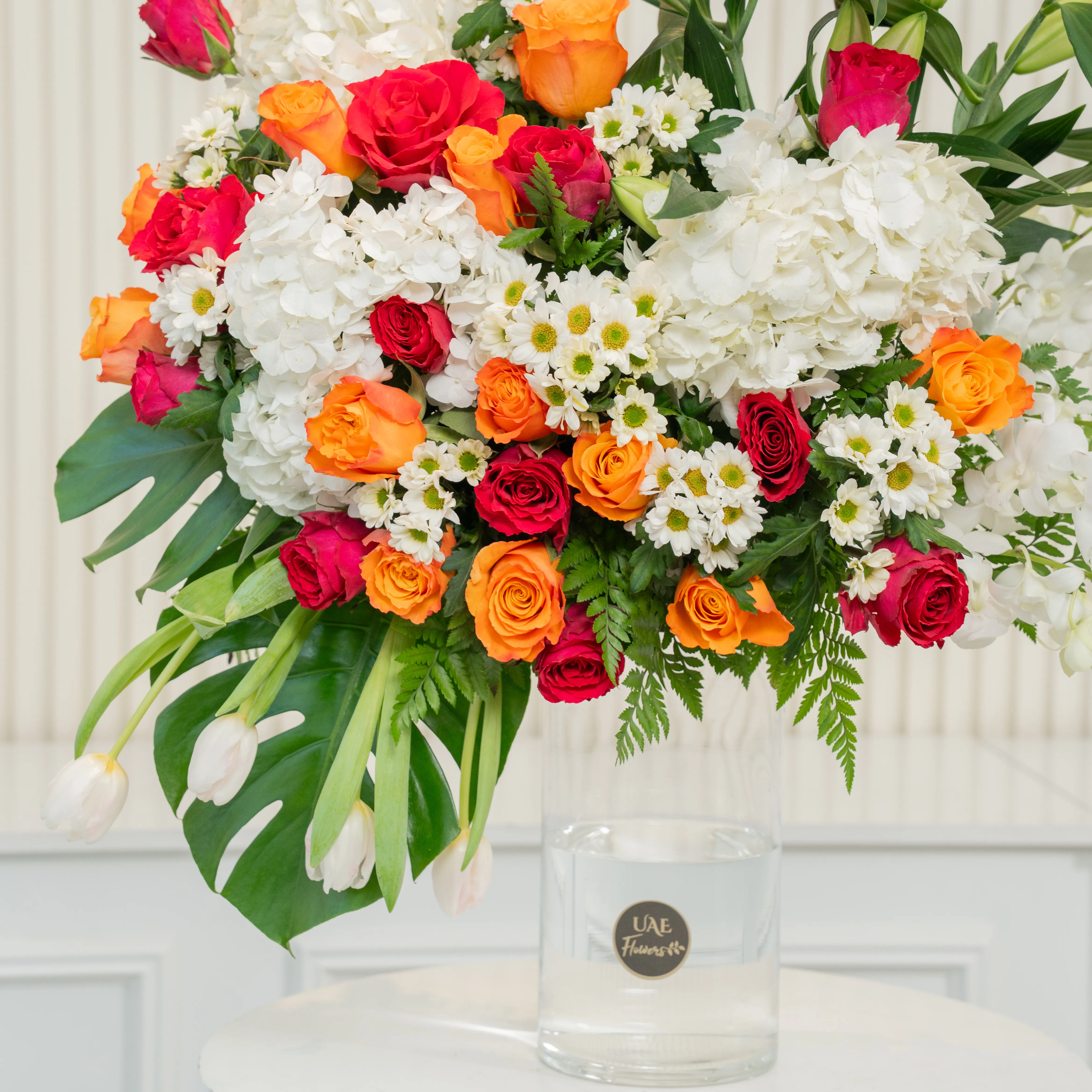 Glass vase arrangement of 3 white lilies, 3 White Hydrangeas, 7 White Orchids, 4 White Tulips, 25 Orange and Red Roses, 6 white Chrysanthemums with green fillers.