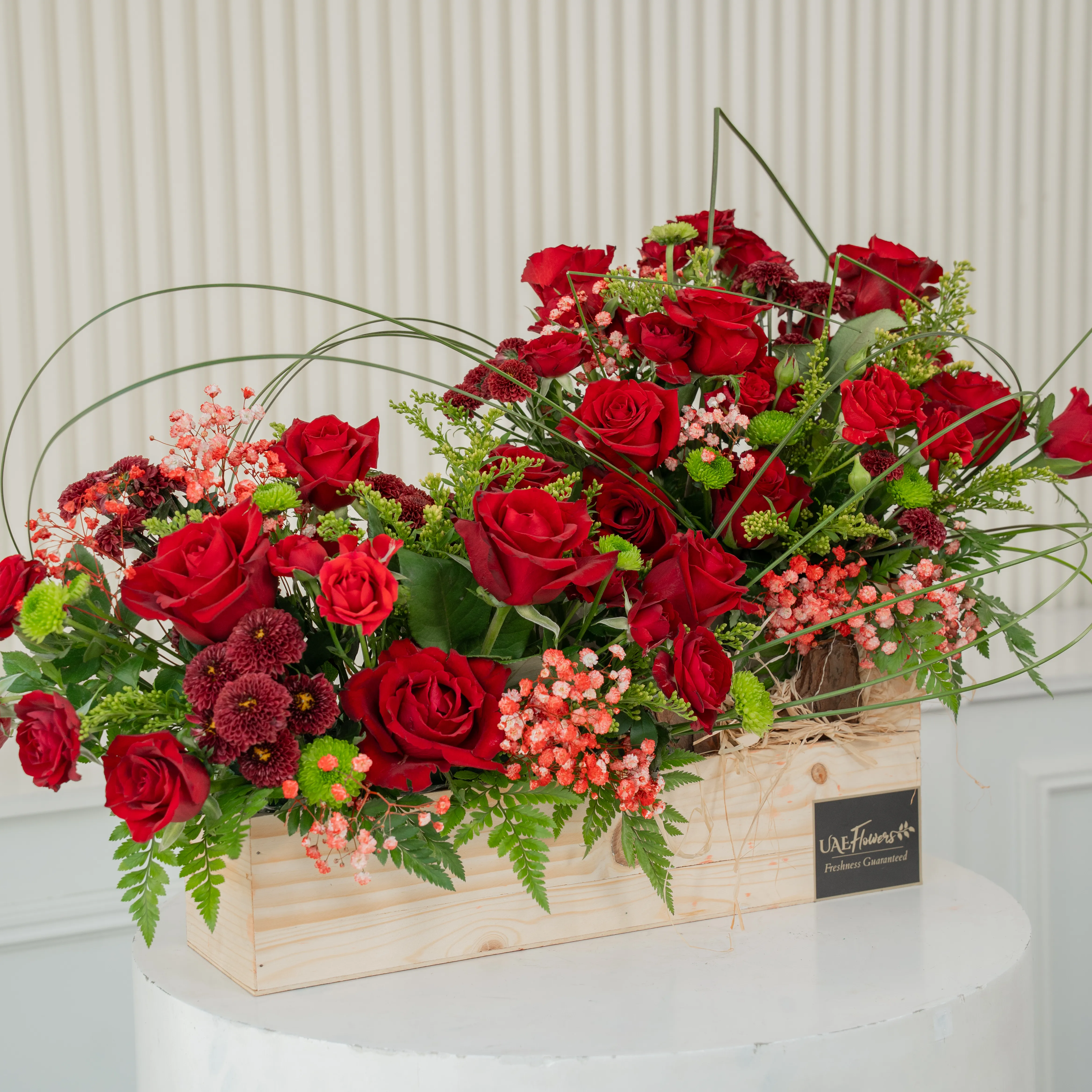 An arrangement of 15 red roses, 7 red spray roses, 4 maroon button chrysanthemums and 4 green button chrysanthemums and steel grass with green fillers in a wooden tray.