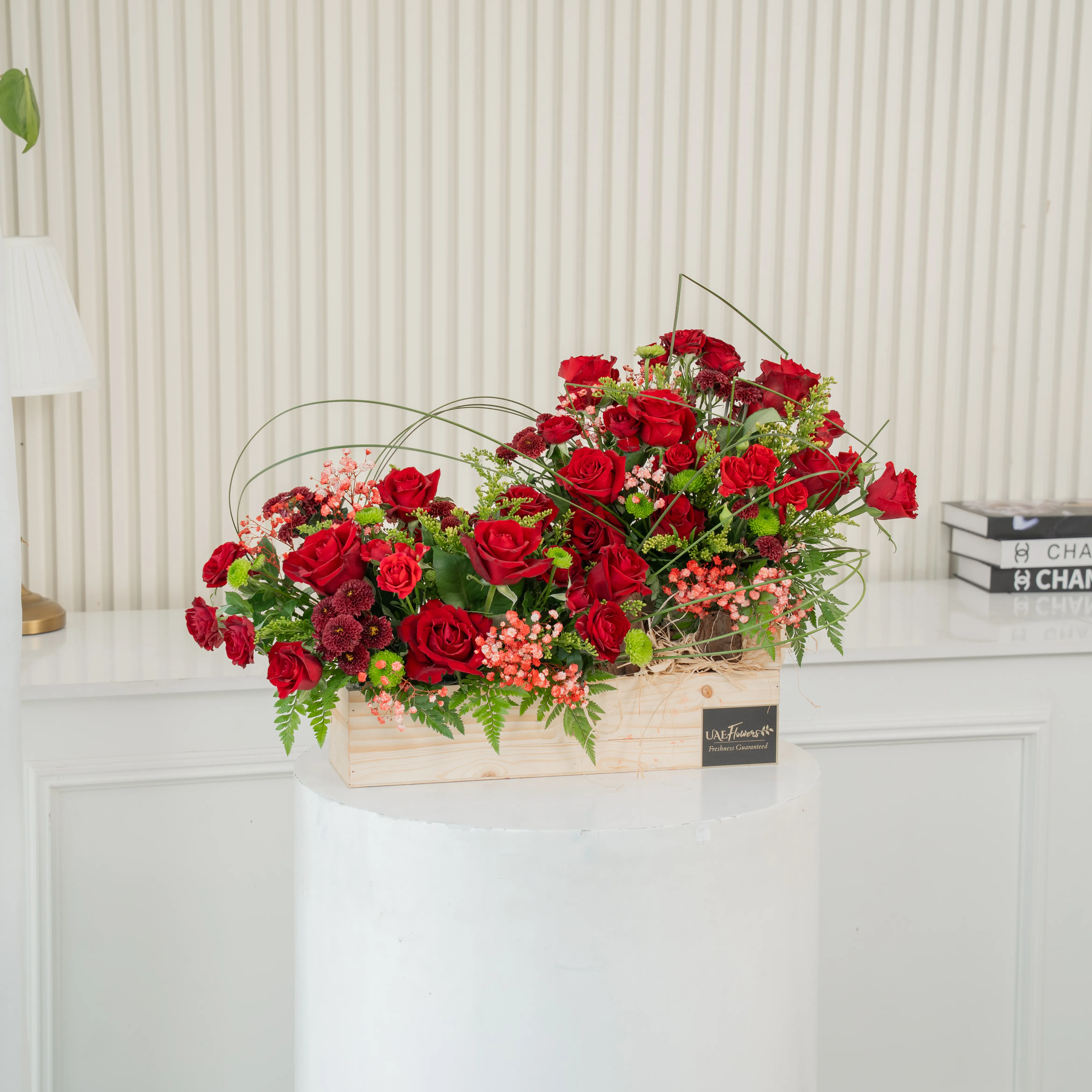 An arrangement of 15 red roses, 7 red spray roses, 4 maroon button chrysanthemums and 4 green button chrysanthemums and steel grass with green fillers in a wooden tray.