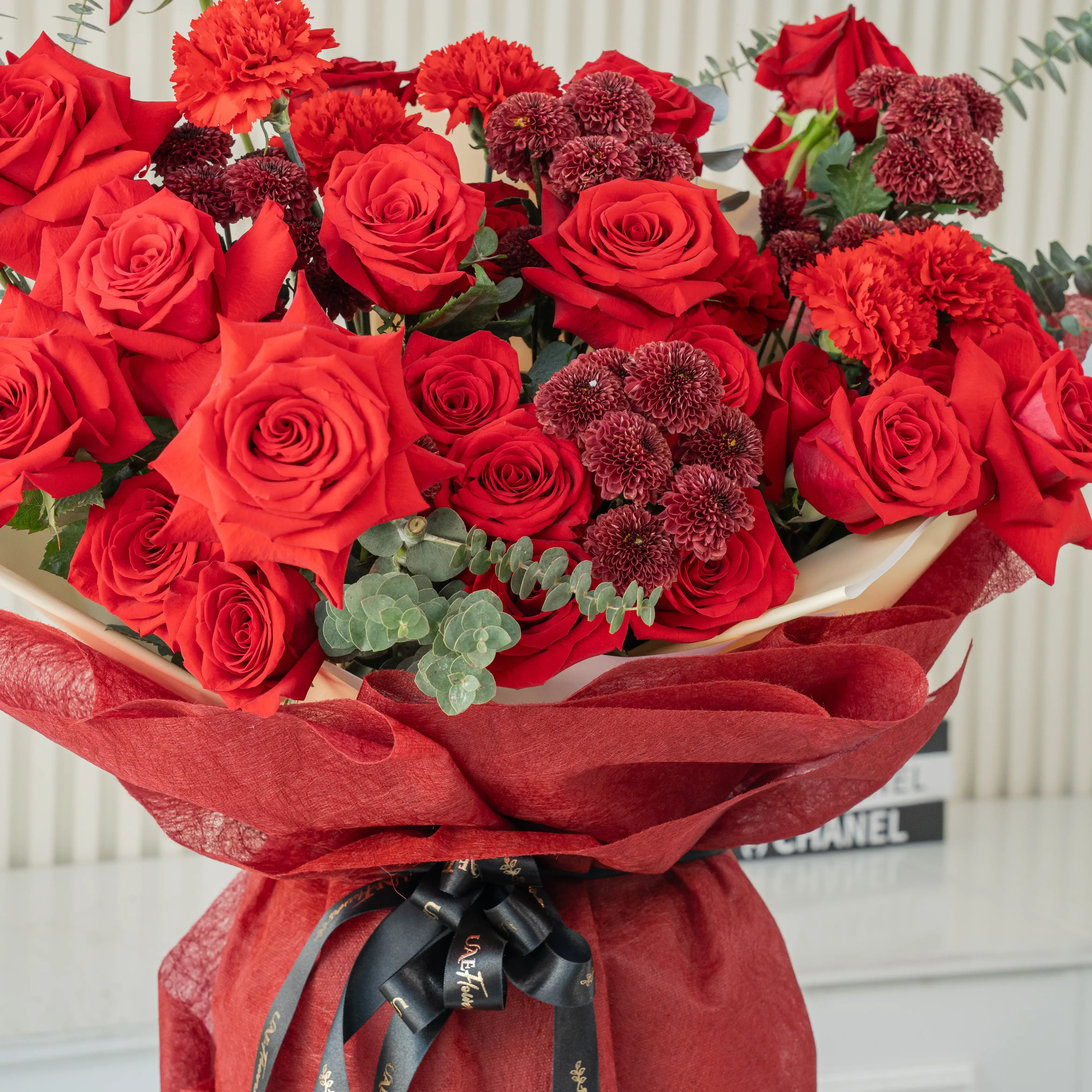 Bouquet of 10 red roses, 5 red spray roses and 3 red button chrysanthemums with eucalyptus leaf in red paper packing tied with black ribbon.