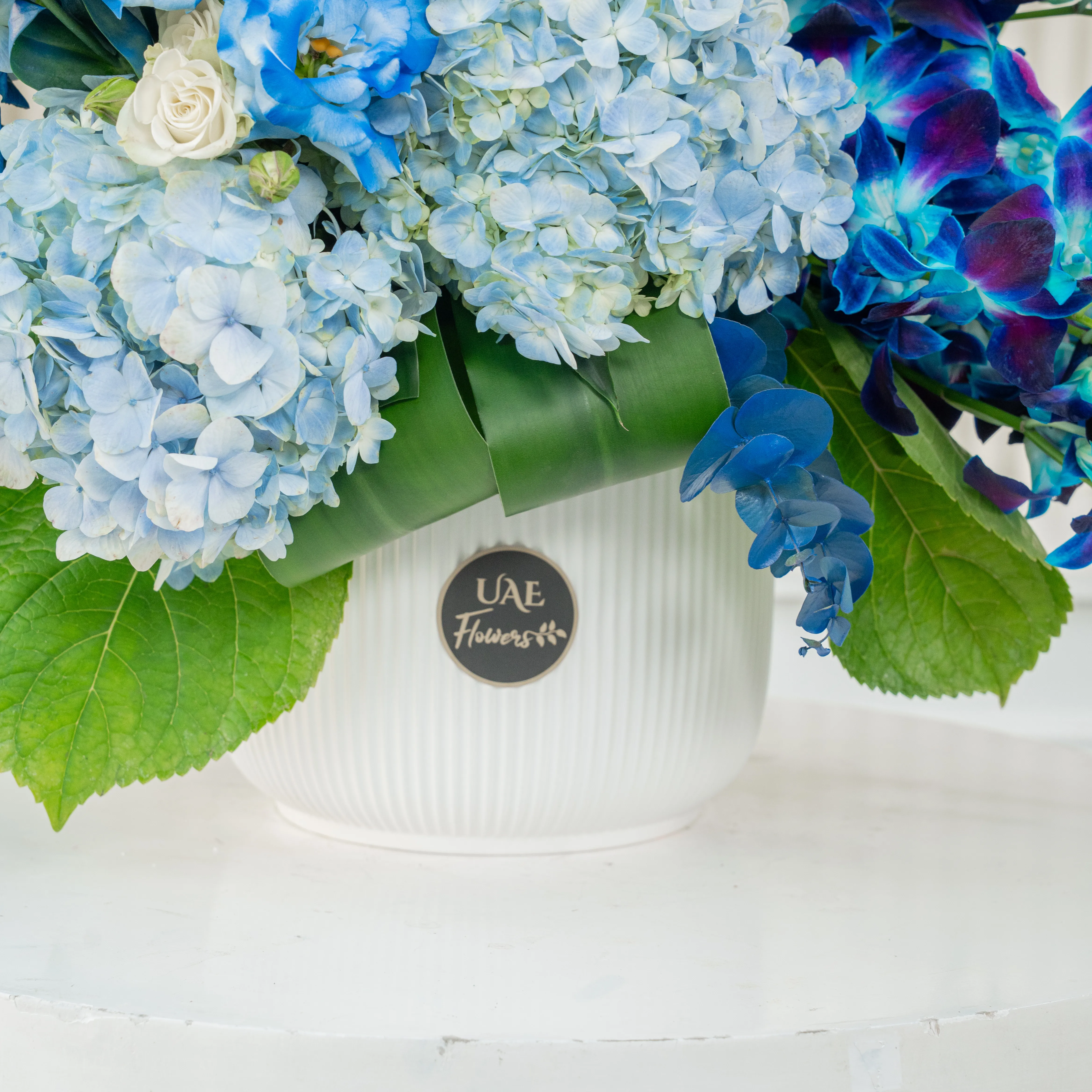 An arrangement of 10 white lisinathus, 5 white matthiola, 10 blue orchids, 5 white roses, 10 white spray roses and 3 blue hydrangeas with eucalyptus leaf and Limonium fillers in a ceramic pot.