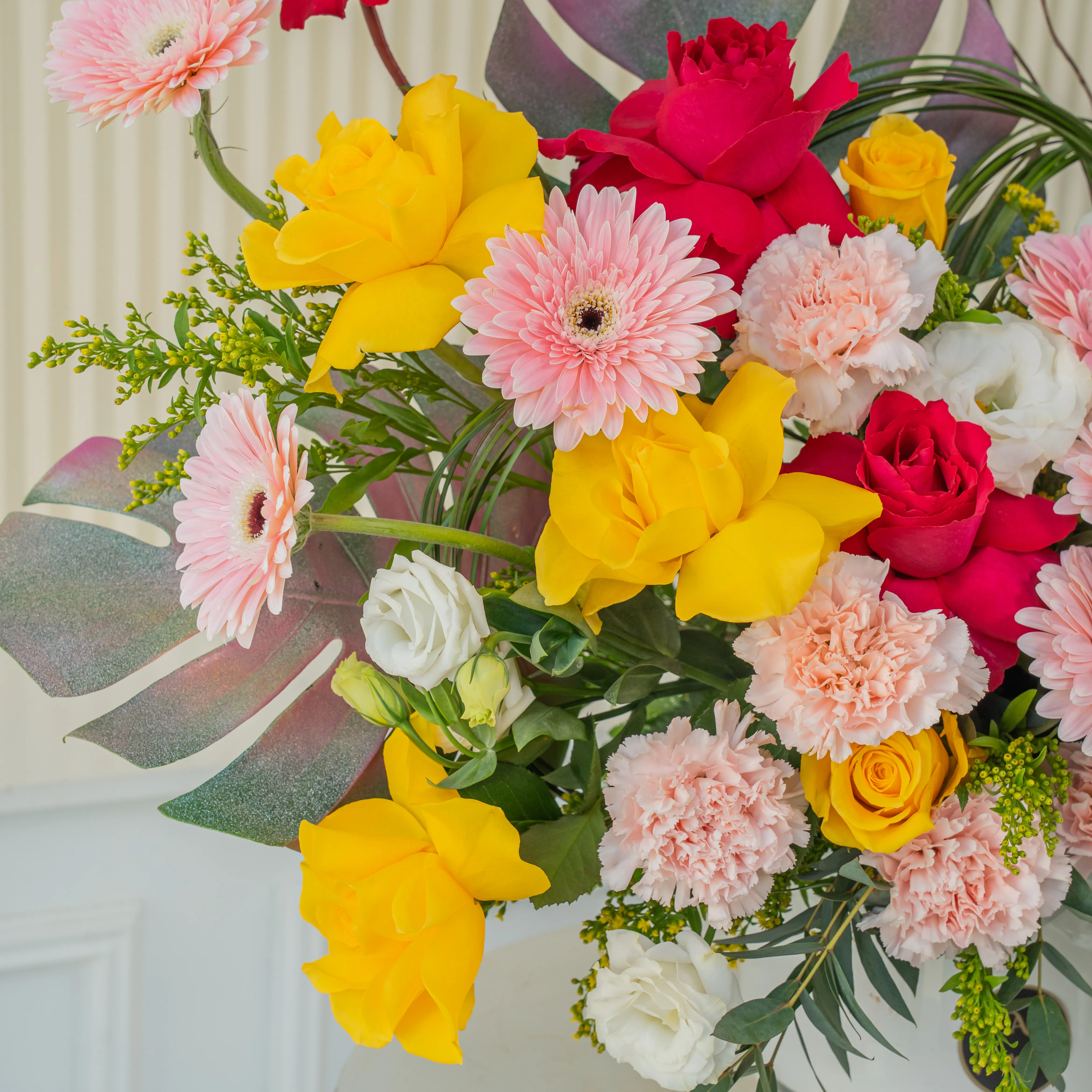 An arrangement of 10 yellow roses, 5 fuchsia roses, 10 pink gerberas, 12 pink carnations, 4 white lisianthus, 4 white orchids with green fillers in a ceramic pot.