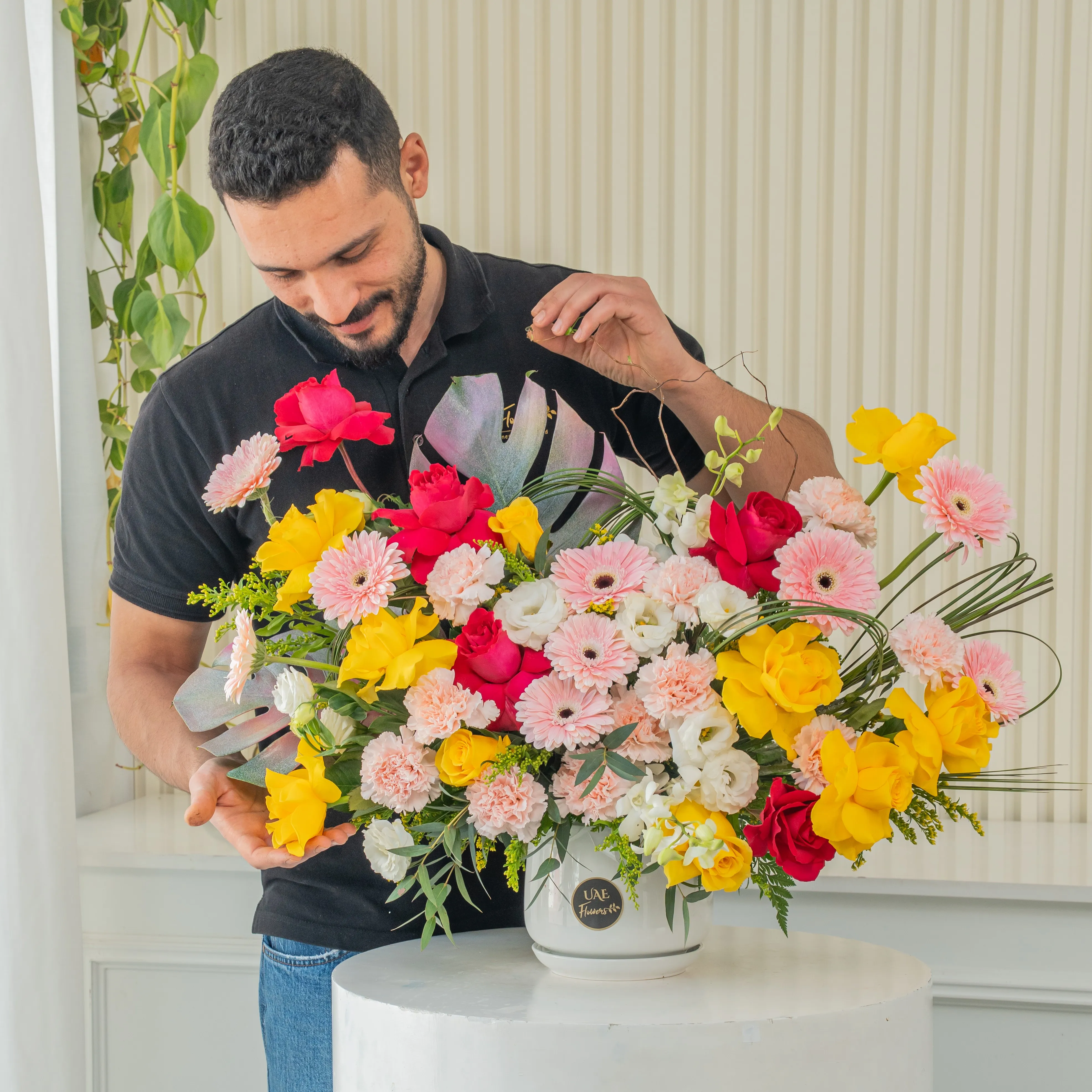 An arrangement of 10 yellow roses, 5 fuchsia roses, 10 pink gerberas, 12 pink carnations, 4 white lisianthus, 4 white orchids with green fillers in a ceramic pot.