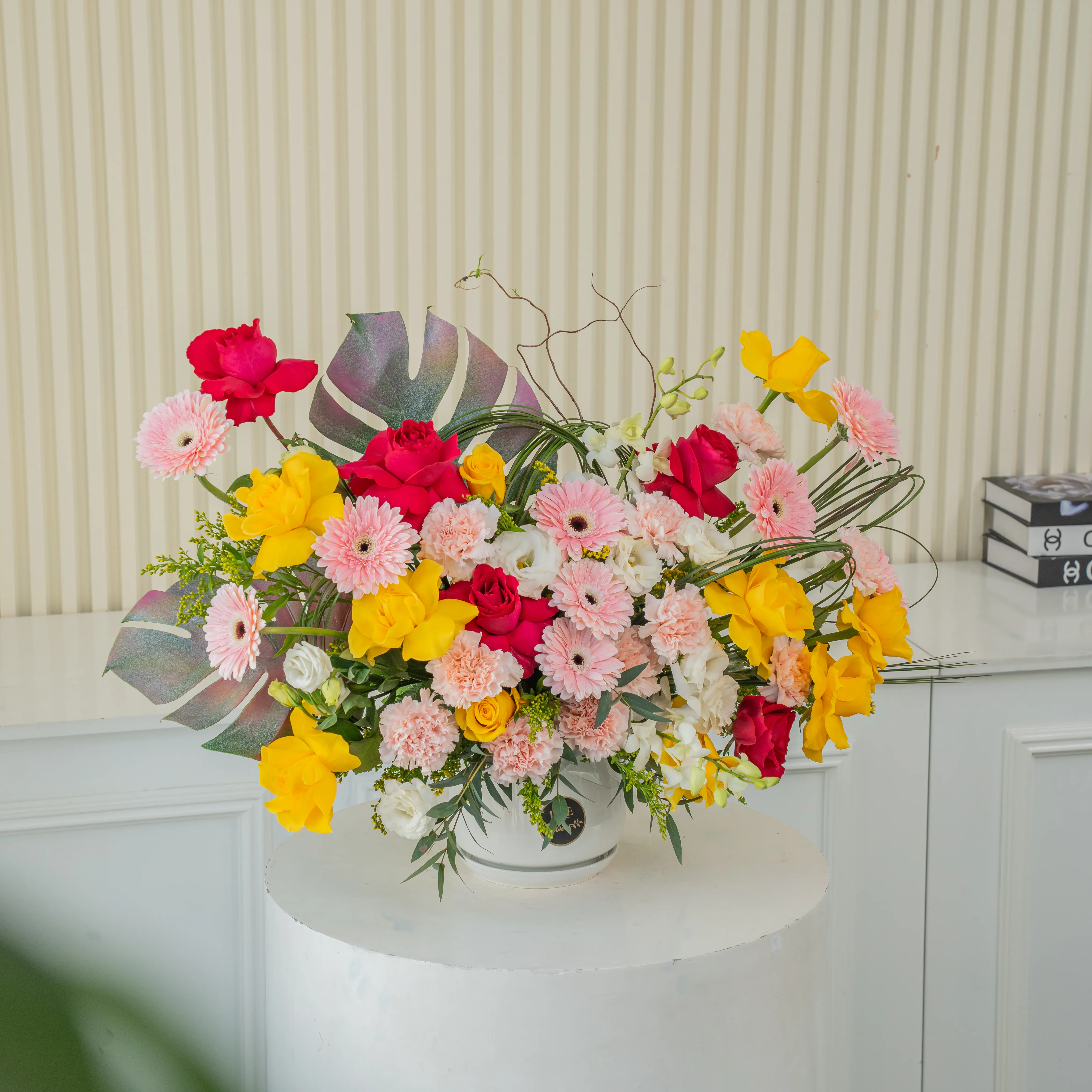 An arrangement of 10 yellow roses, 5 fuchsia roses, 10 pink gerberas, 12 pink carnations, 4 white lisianthus, 4 white orchids with green fillers in a ceramic pot.