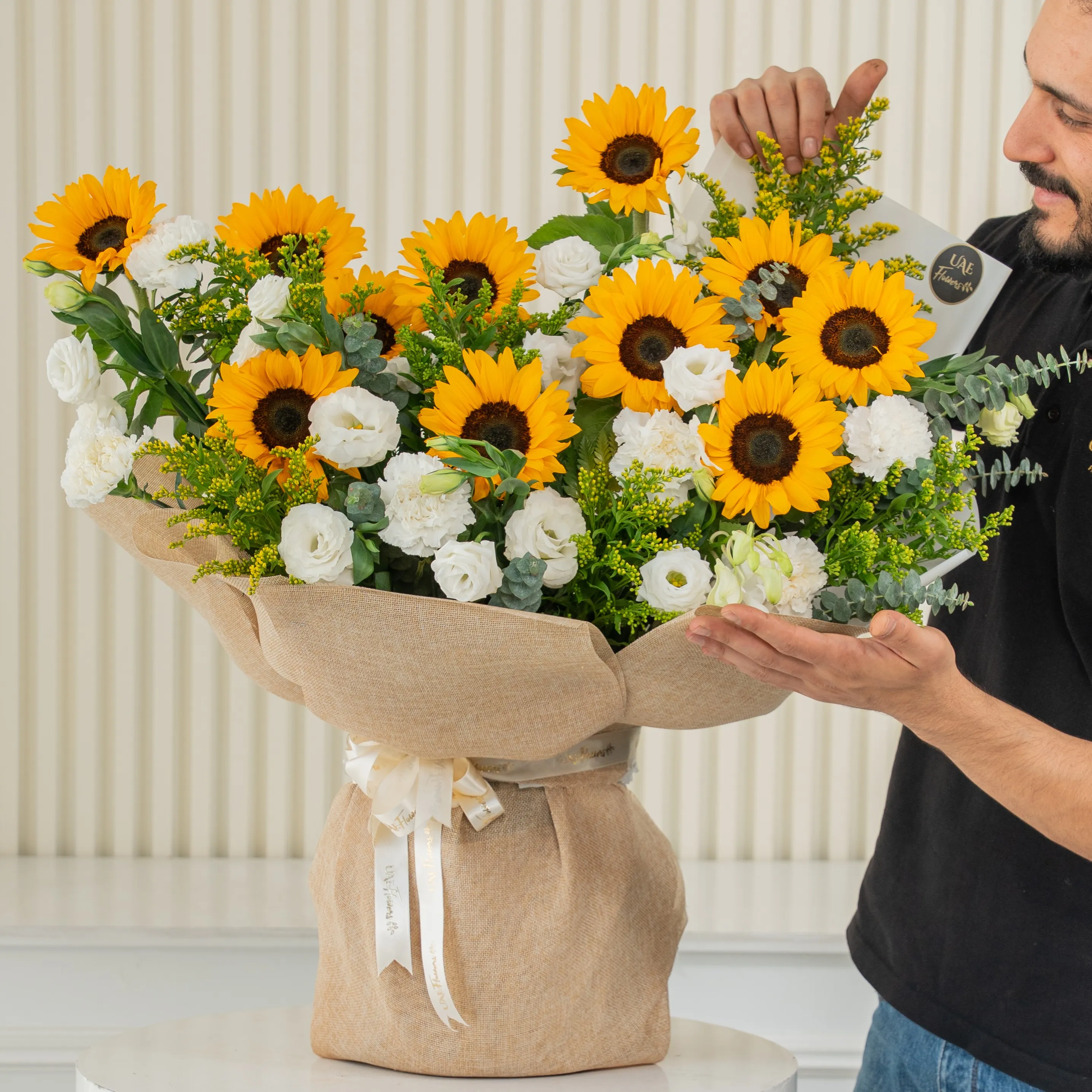 Bouquet of 11 sunflowers and 10 white lisianthus with green fillers and yellow statis in brown paper packing tied with white ribbon.